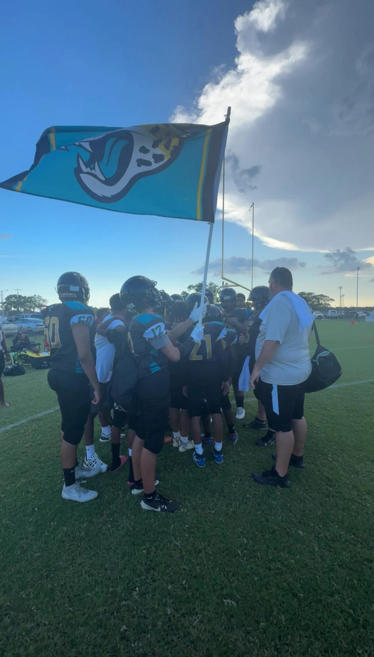 Football team huddled on a field with coach, one player holding a team flag, under a partly cloudy sky.