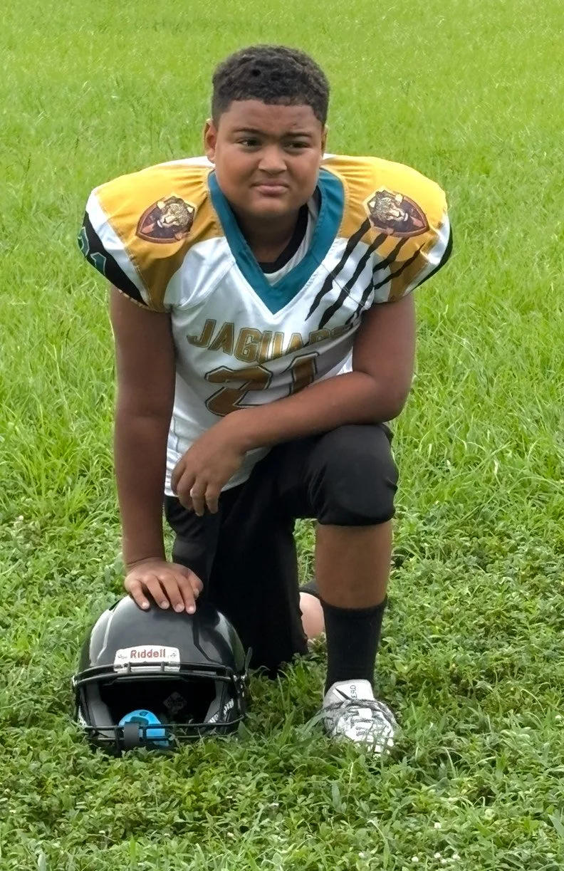Young football player in uniform kneeling on grass with helmet on the ground.