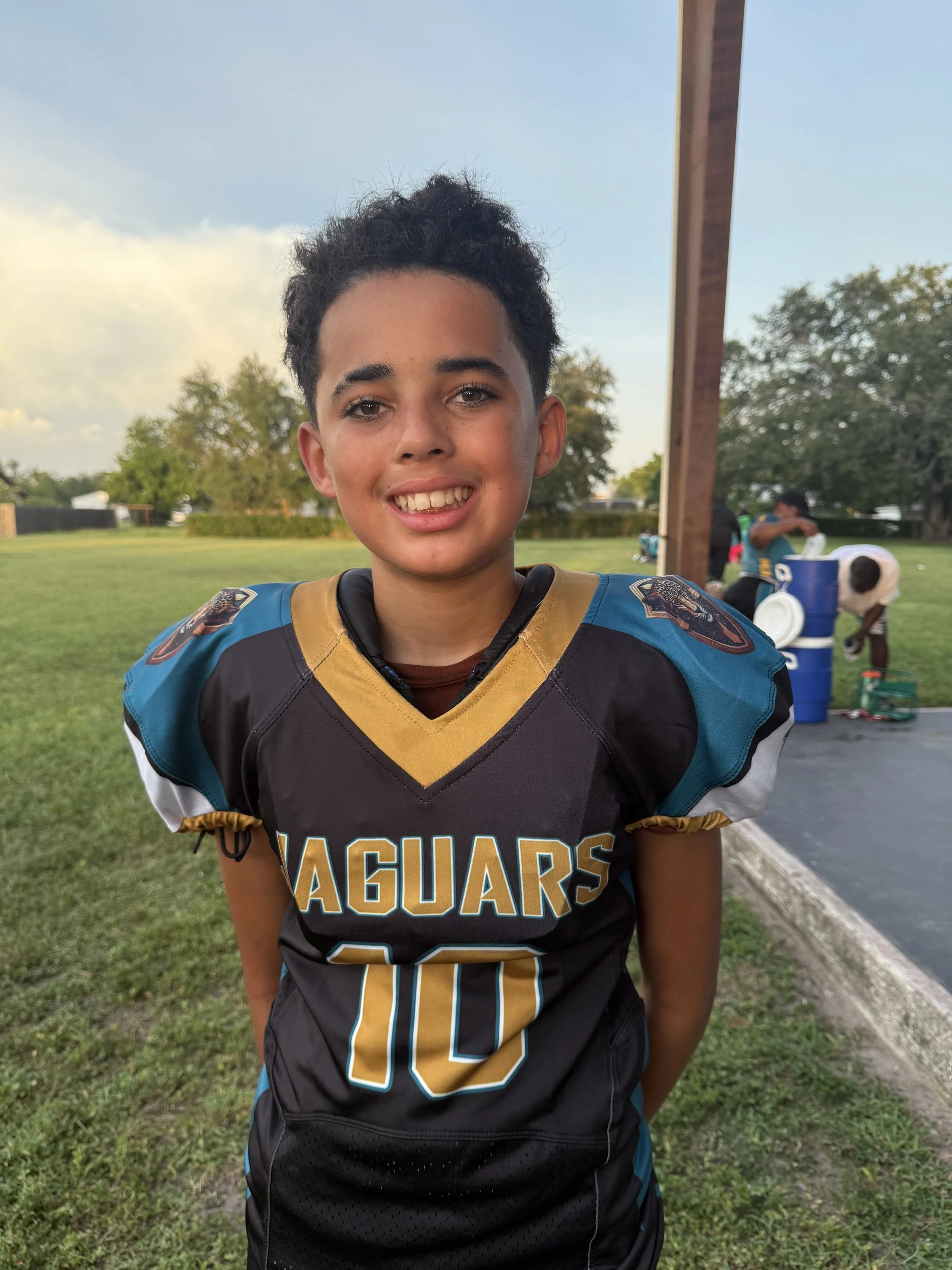 A young boy wearing a black football jersey with teal and gold accents, labeled 'Jaguars' and the number 10, standing outside on a grassy field with a blue sky, trees, and a few people in the background.
