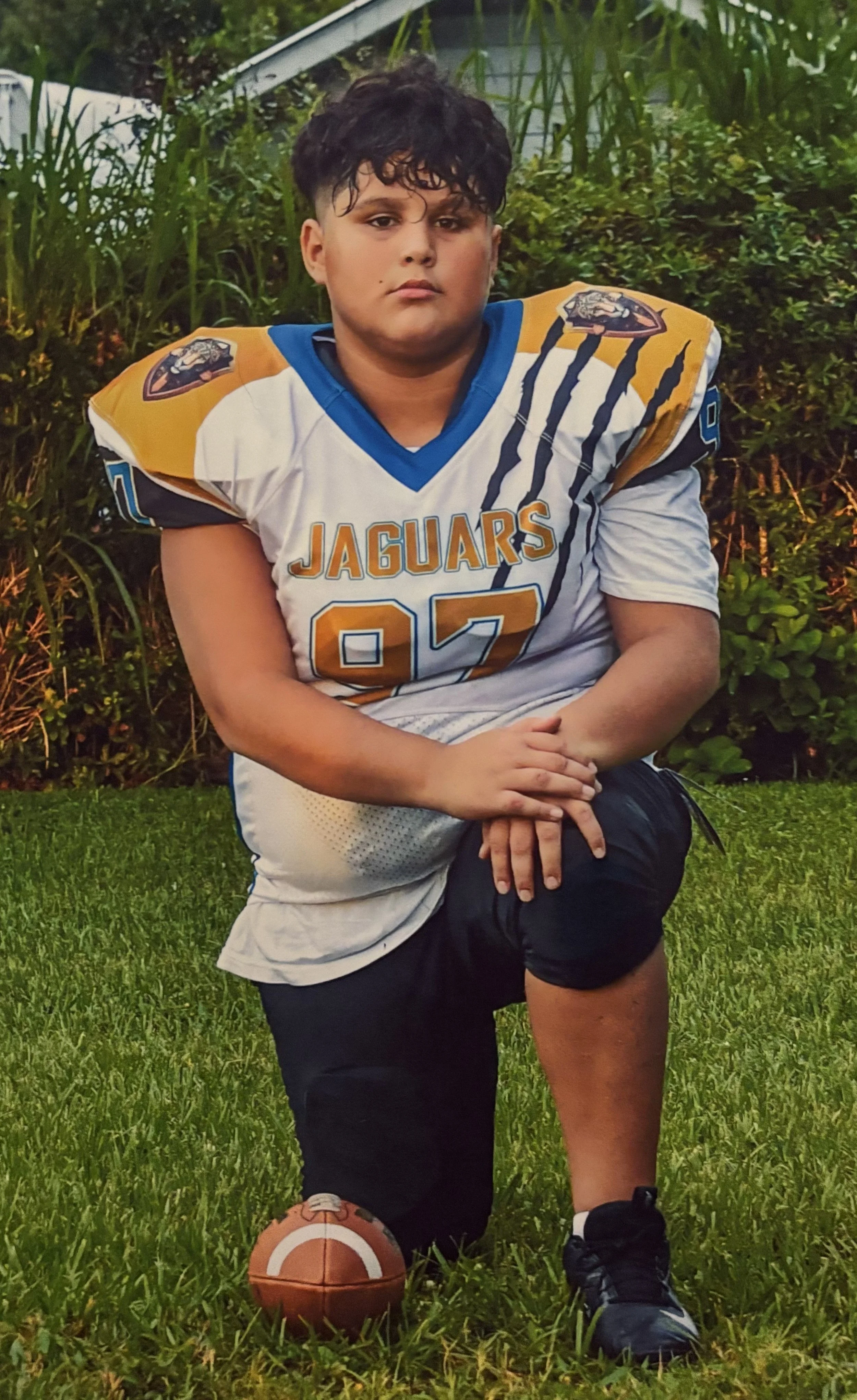 Young football player in a Jaguars uniform kneeling on grass with one knee, an American football on the ground beside him, in front of green bushes.