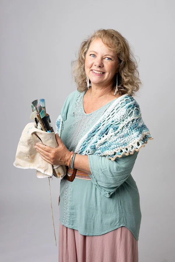 Image of Kariba smiling with her paint brushes bundled in her hands.  She is wearing a hand-made shawl in the color of the ocean.