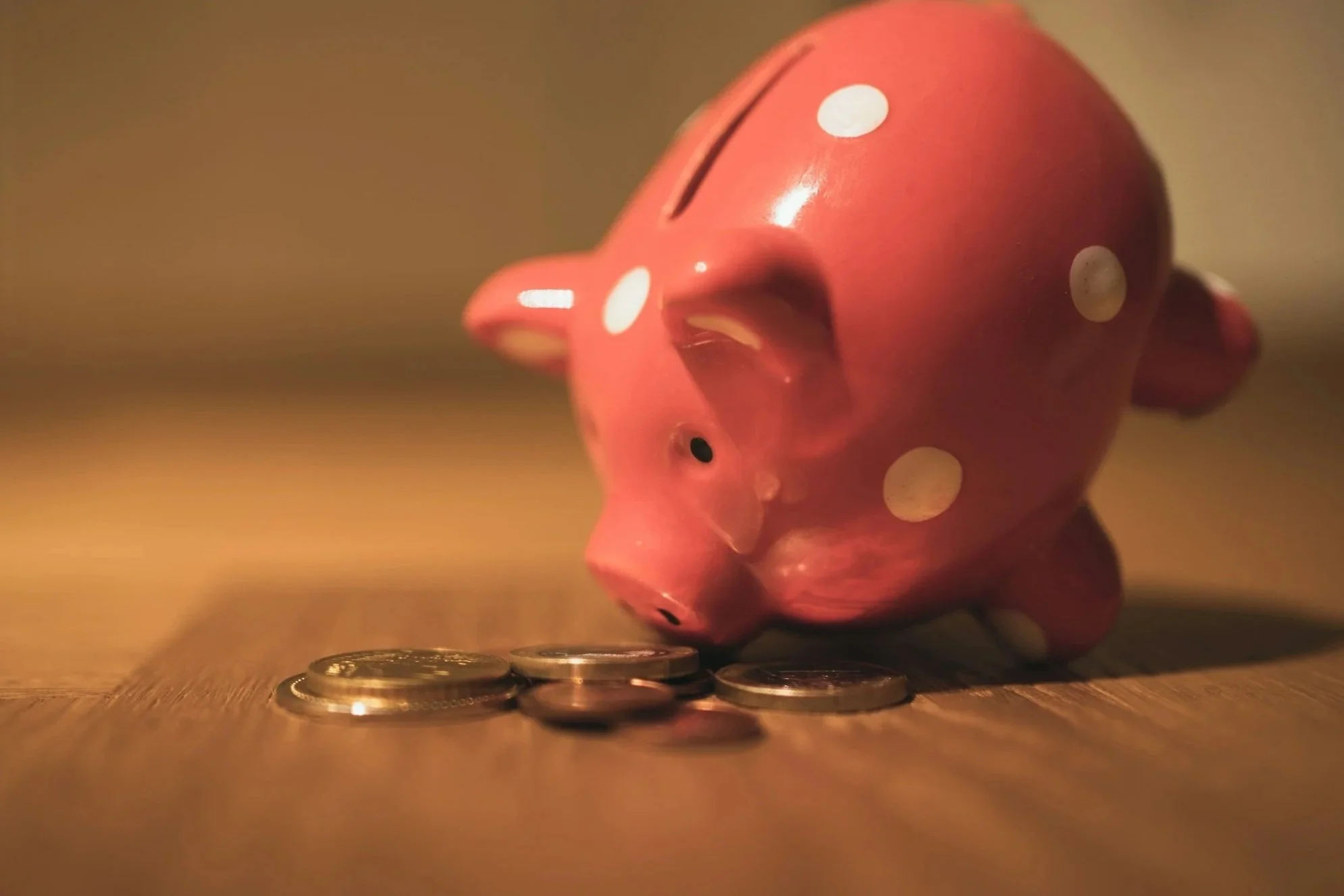 Pink ceramic piggy bank with white polka dots, lying on its side with coins scattered beneath it on a wooden surface, in warm lighting.