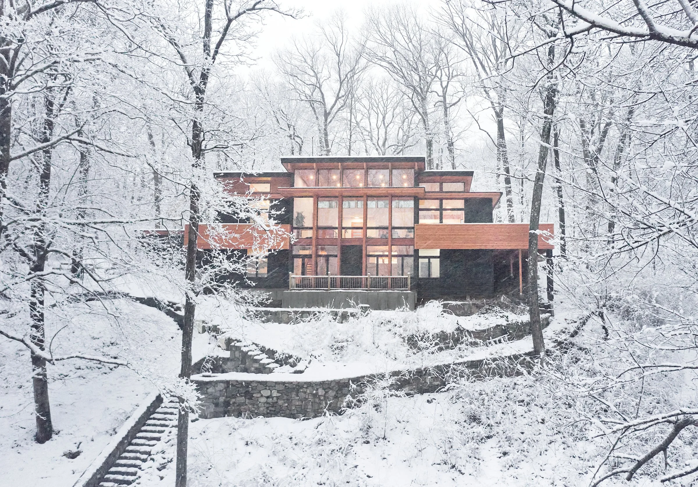Modern house with large glass windows and wooden accents, located on a snow-covered hillside in a forest of leafless trees.