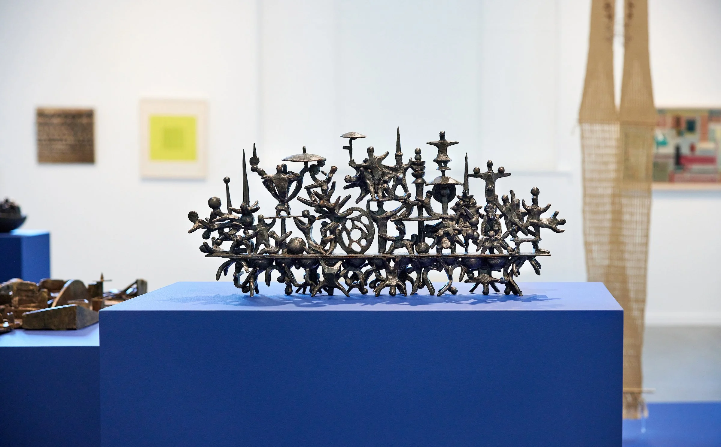 A detailed bronze sculpture featuring numerous small, interconnected human figures, displayed on a blue pedestal in an art gallery.
