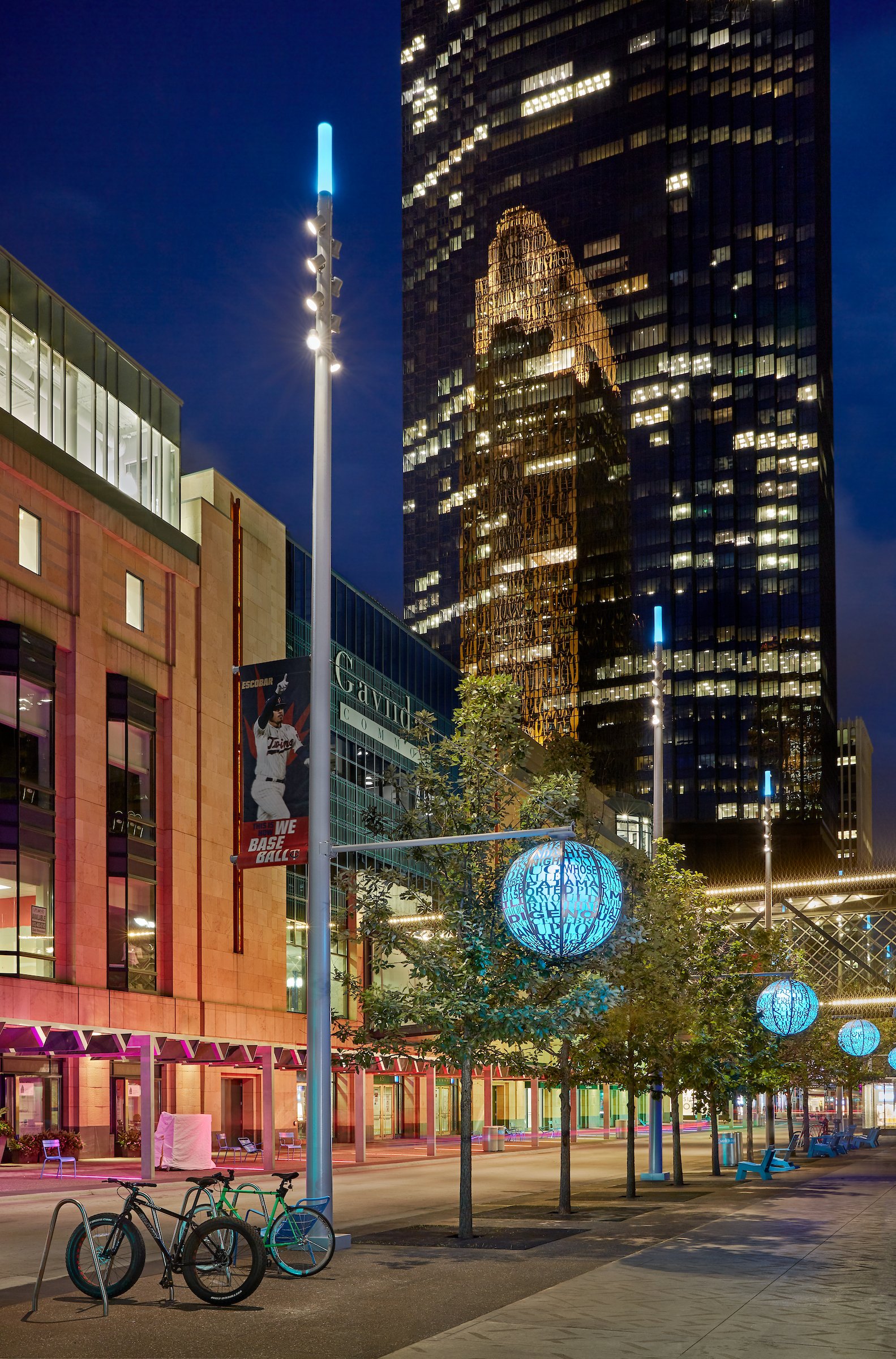 City street at night with illuminated trees, modern buildings, colorful lights, and bicycles parked on sidewalk.