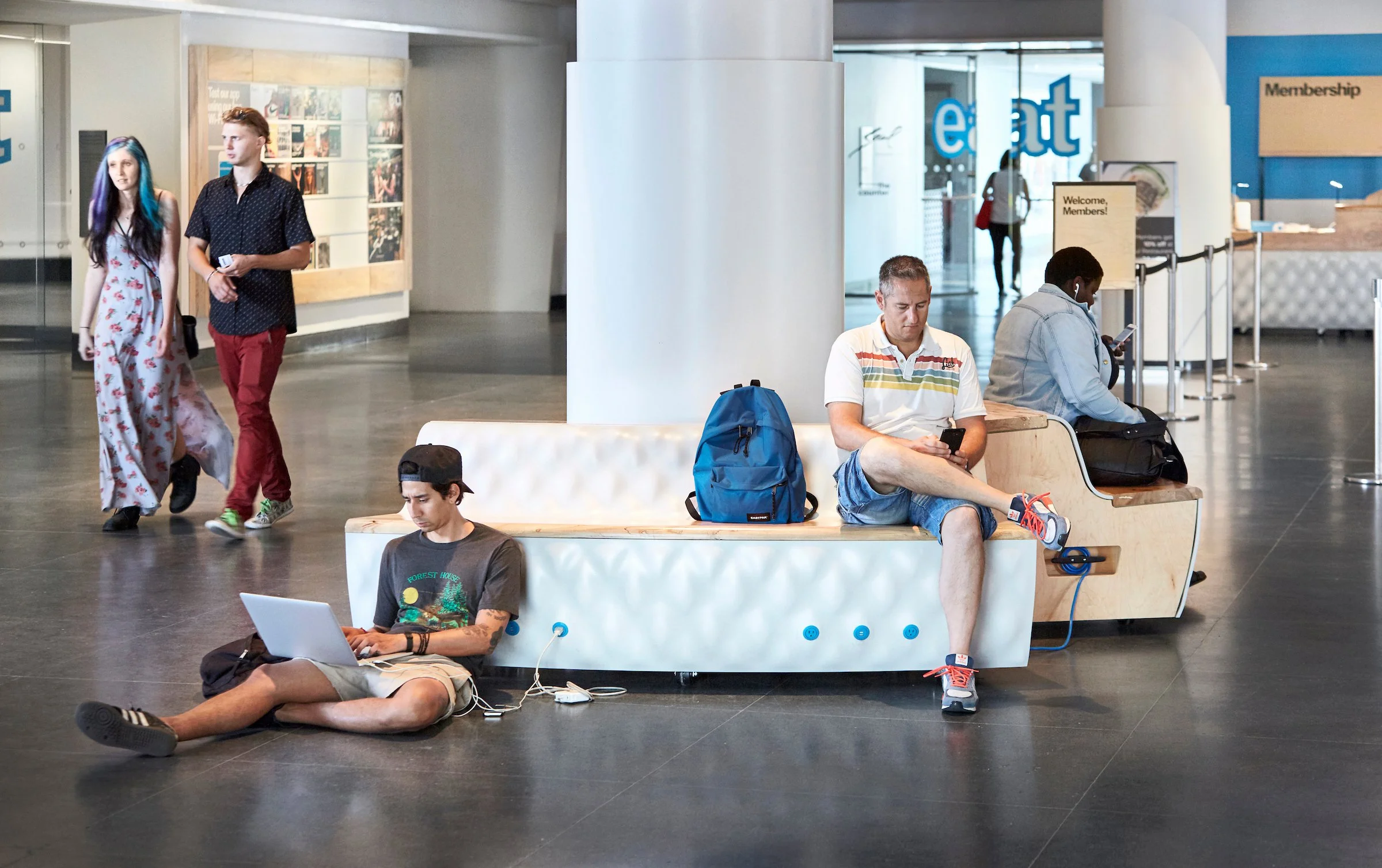 Four people in an airport. Three are sitting on a modern bench with their phones, and one person is sitting on the floor with a laptop. Two other individuals are walking past in the background.