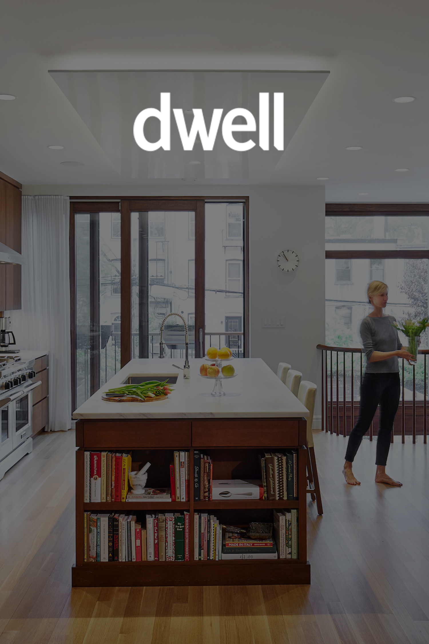 Cover photo of Dwell magazine featuring a modern kitchen with a kitchen island, books on a lower shelf, and a woman carrying a tray of green vegetables.