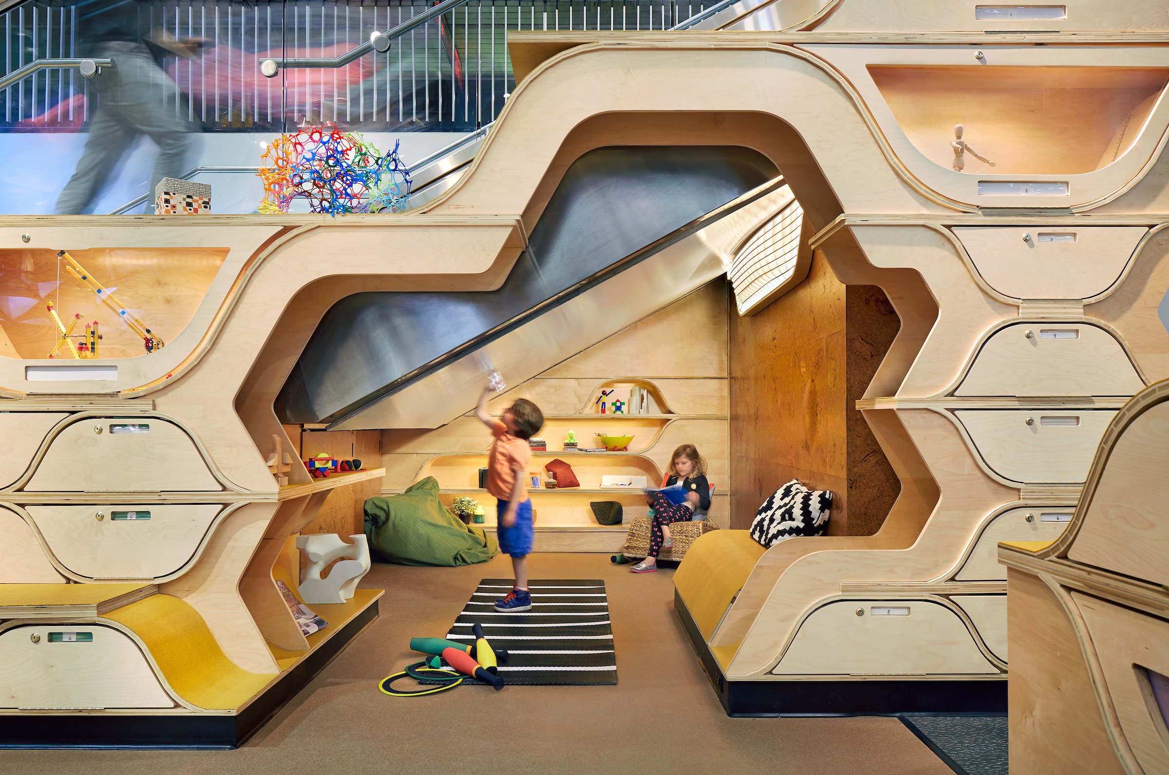 Children playing in a modern indoor playground with wooden structures, a slide, and various toys and activities.