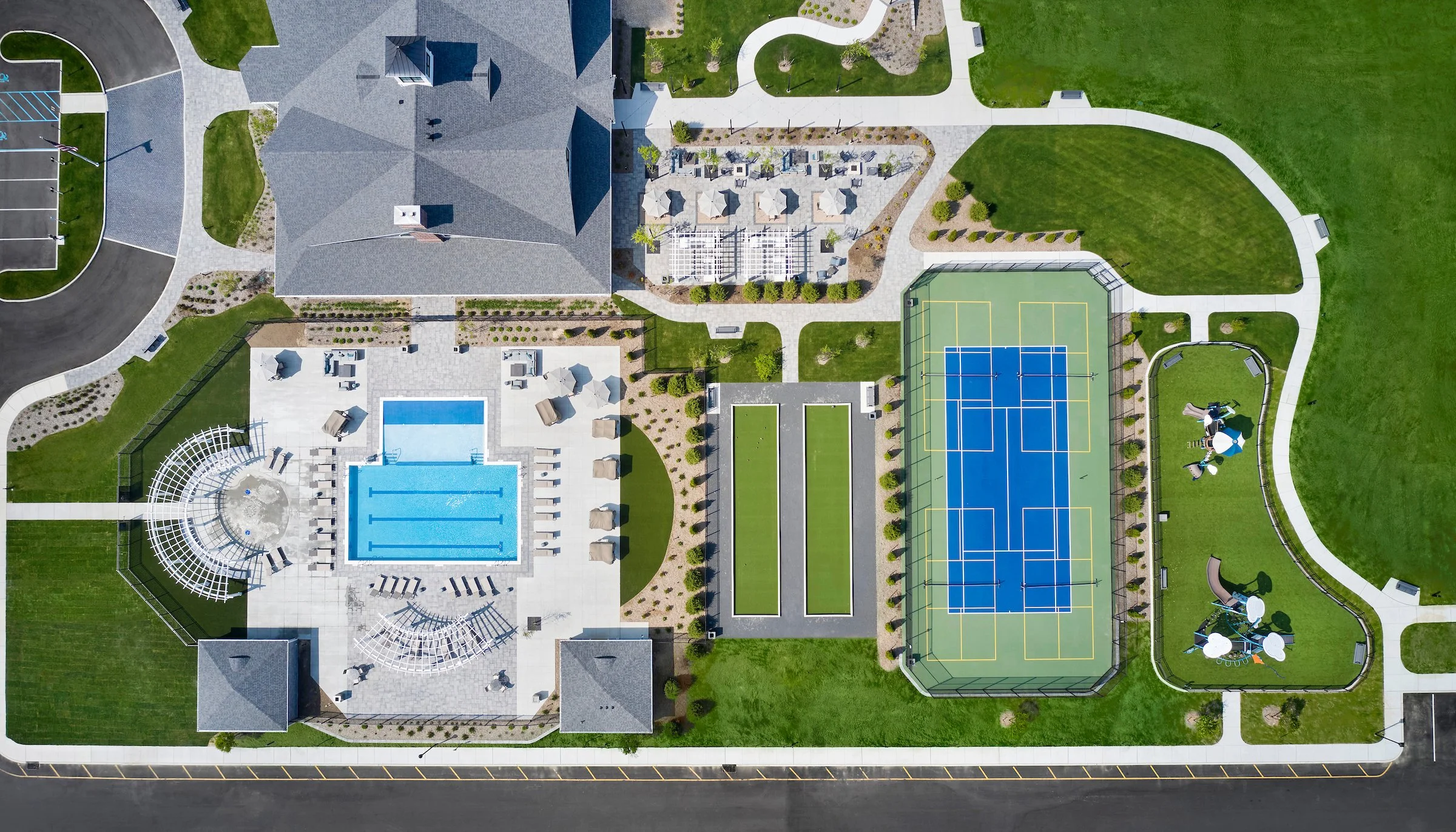 An aerial view of a recreational complex featuring a swimming pool, tennis courts, a playground, and green lawns, surrounded by walking paths and parking areas.