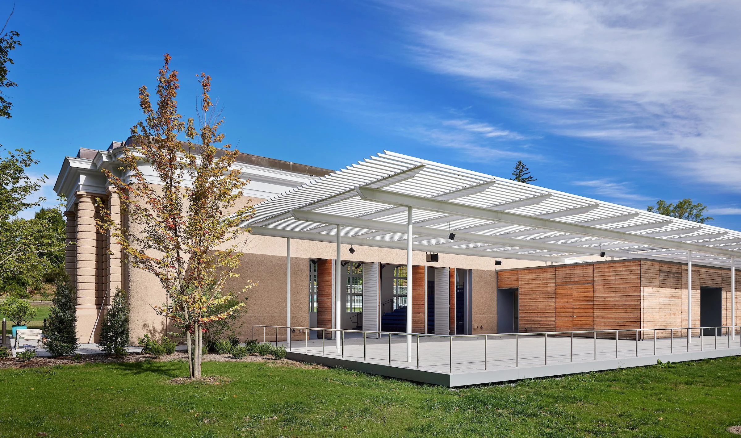 Modern building with beige and wood exterior, white metal pergola, landscaped lawn, trees, and a blue sky.