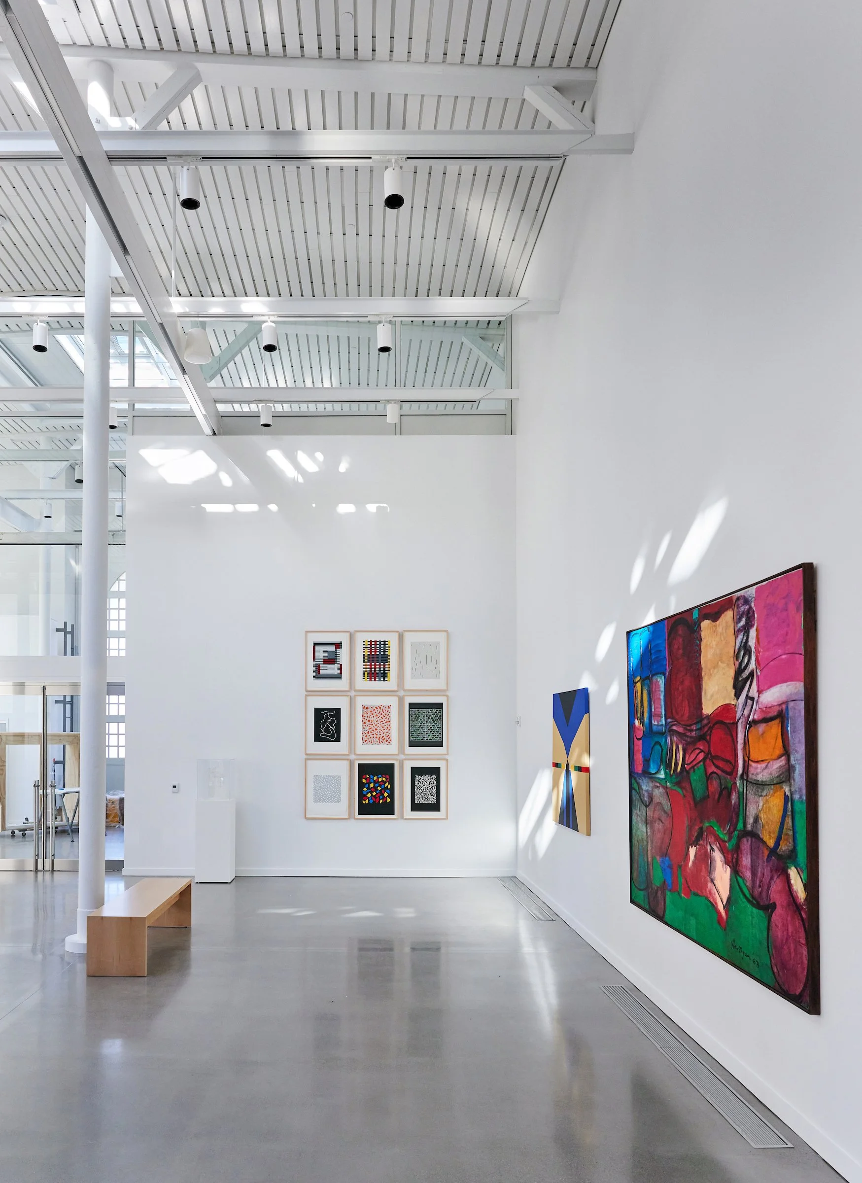 An art gallery with white walls, featuring various abstract paintings and artworks, with a bench on the floor and sunlight streaming through the ceiling.