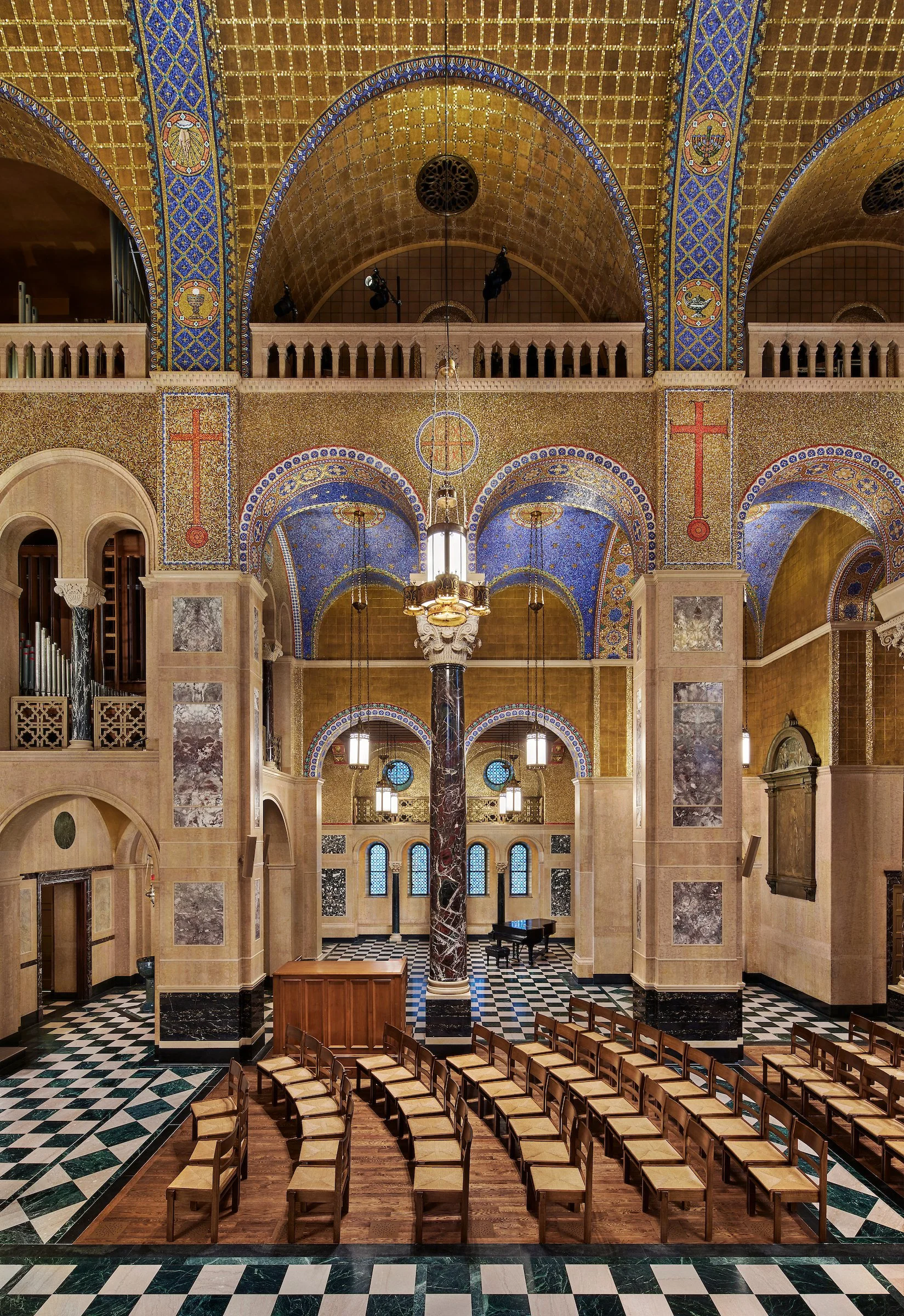Interior of an ornate church or cathedral with gold mosaic ceilings, arched ceilings, and a central column. Wooden chairs are arranged in rows facing the altar, with a grand piano in the background. The floor has a black and white checkered pattern, 