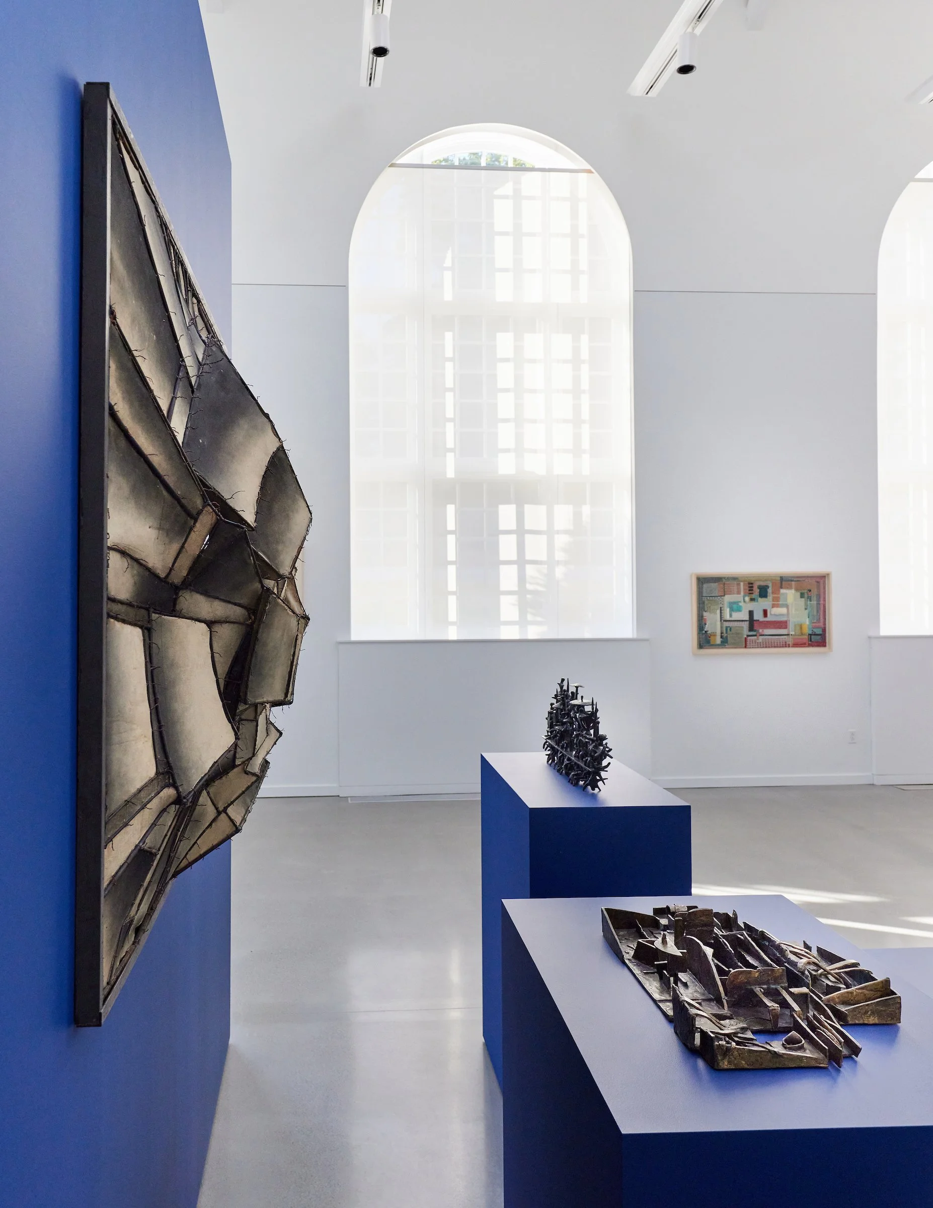 Art gallery with several sculptures and paintings on display, white walls, large arched window, and blue display pedestals.