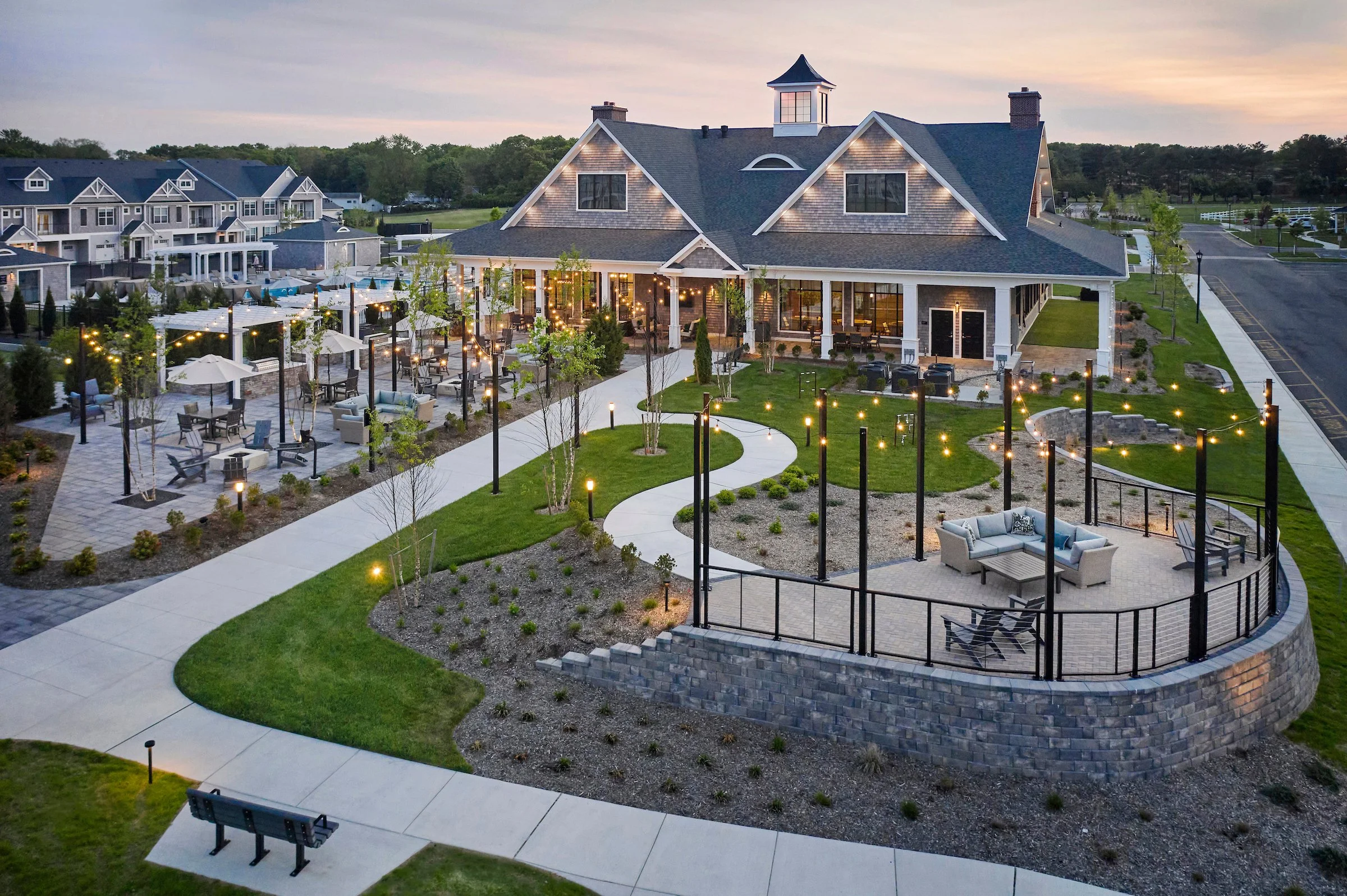A large modern clubhouse with an outdoor lounge area featuring string lights, surrounded by landscaped gardens, walking paths, and additional seating areas at sunset.