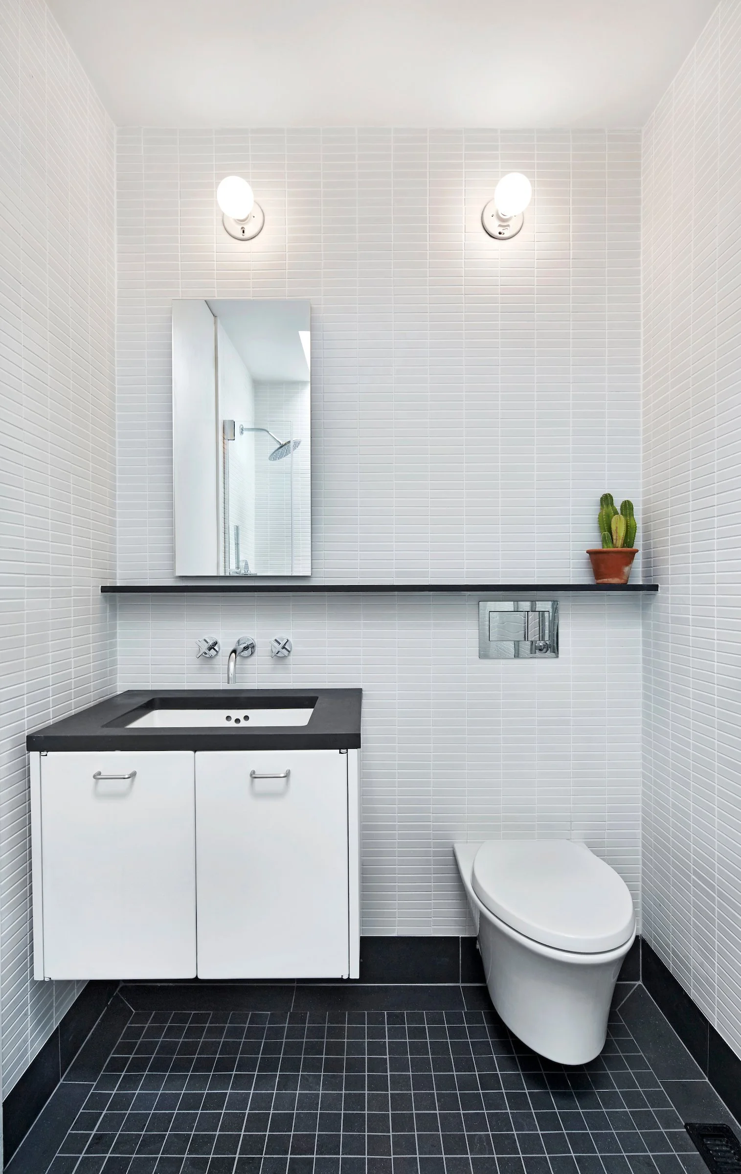 Modern bathroom with white tiled walls, black tiled floor, a white vanity with sink, mirror, cactus plant, and toilet.