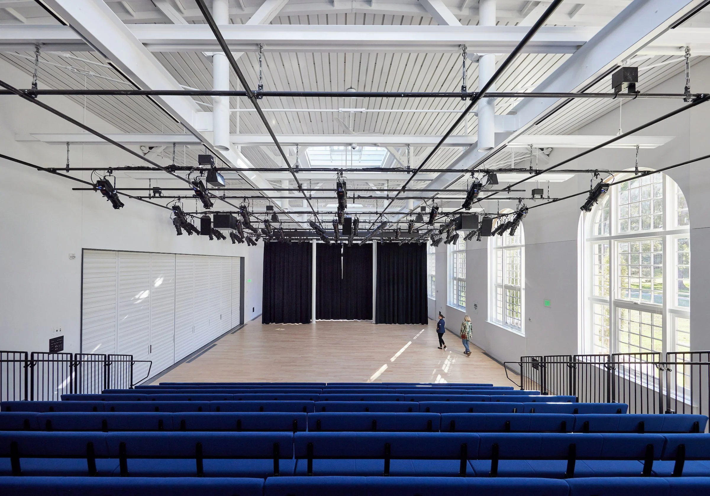 Empty theater or performance hall with a stage, black curtains, rows of blue seats, large windows, and stage lighting equipment hanging from the ceiling.