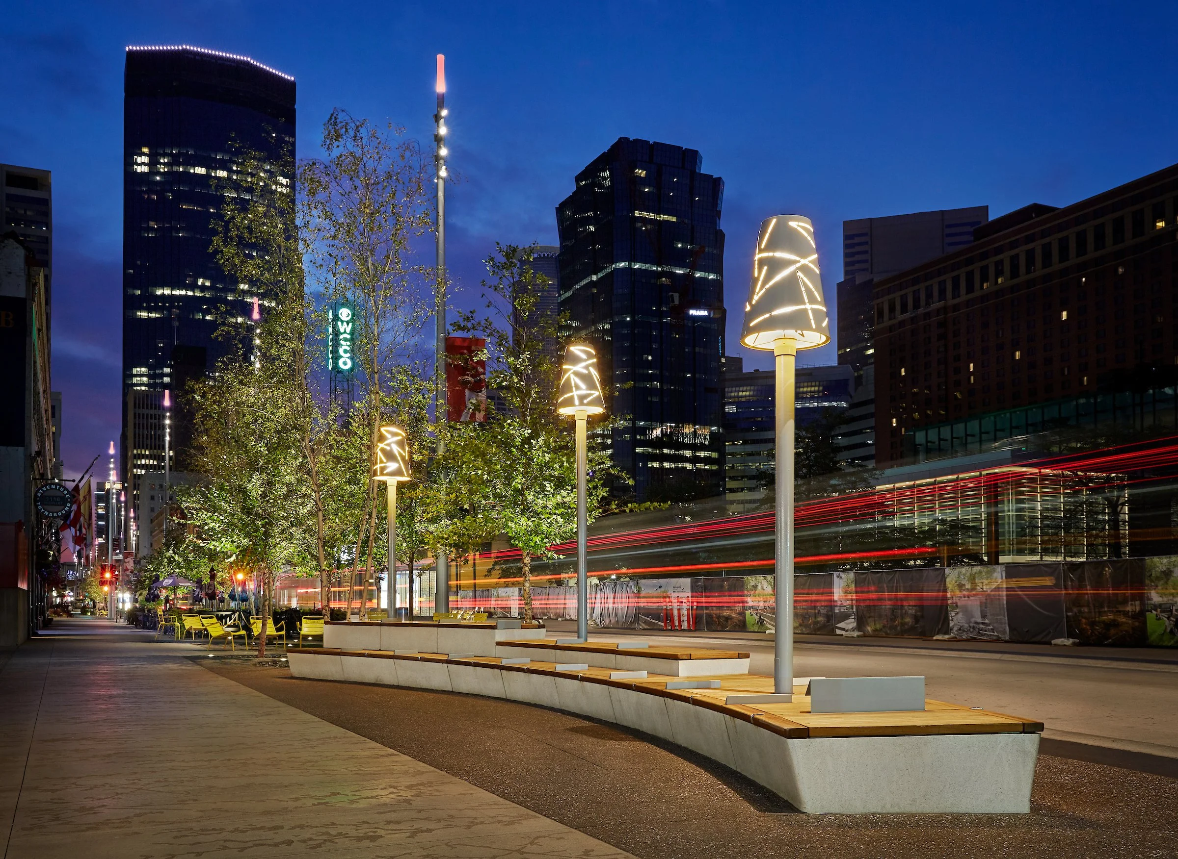 City street at night with illuminated lamp posts, trees, and modern skyscrapers in the background.