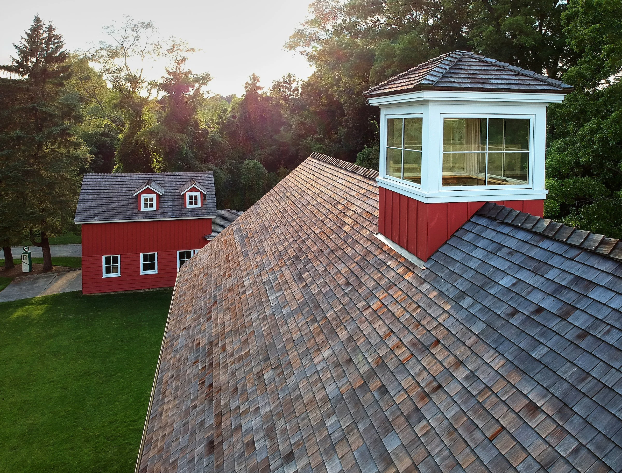A red barn with a small white dormer window in the front and a larger building in the background, both with gray shingle roofs, surrounded by green trees and grass at sunset.