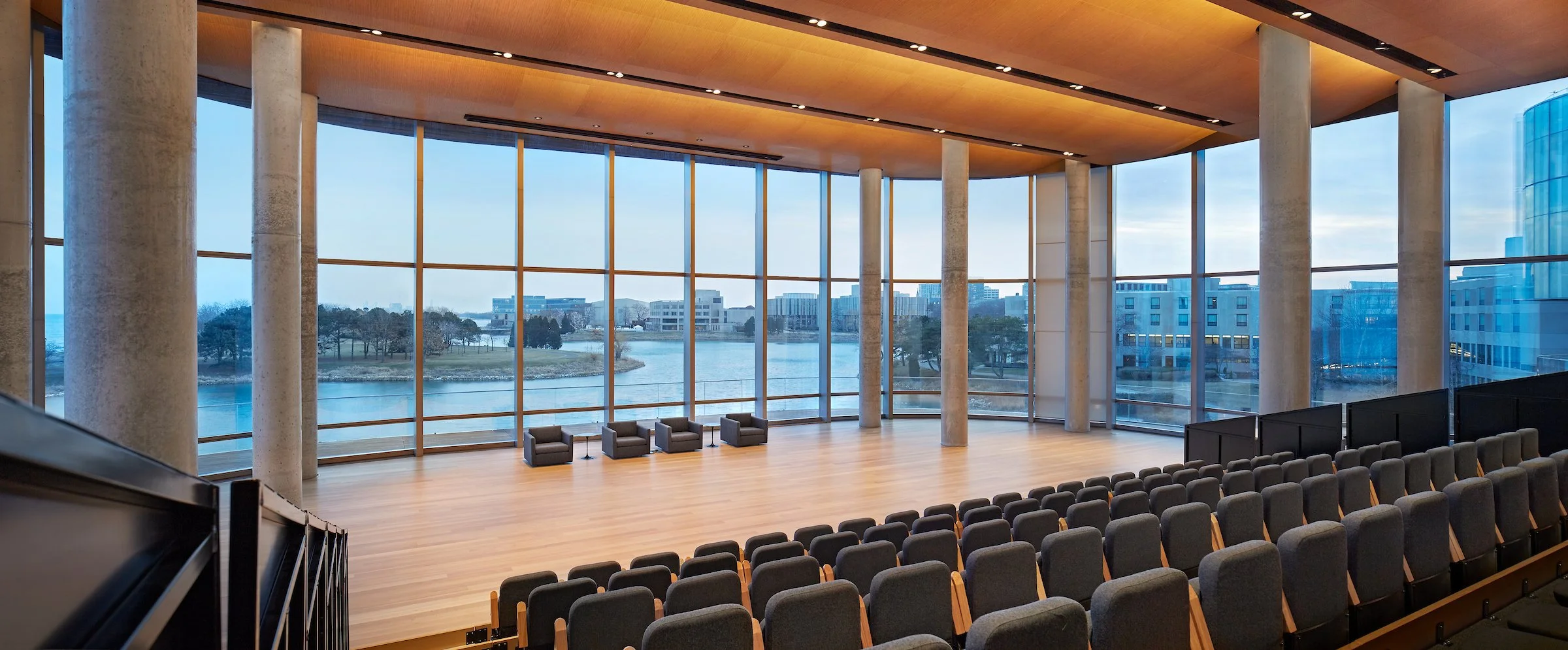 Large modern conference room or auditorium with floor-to-ceiling windows showing an outdoor water view, row of black chairs, four gray armchairs near the window, and a wooden ceiling with recessed lighting.