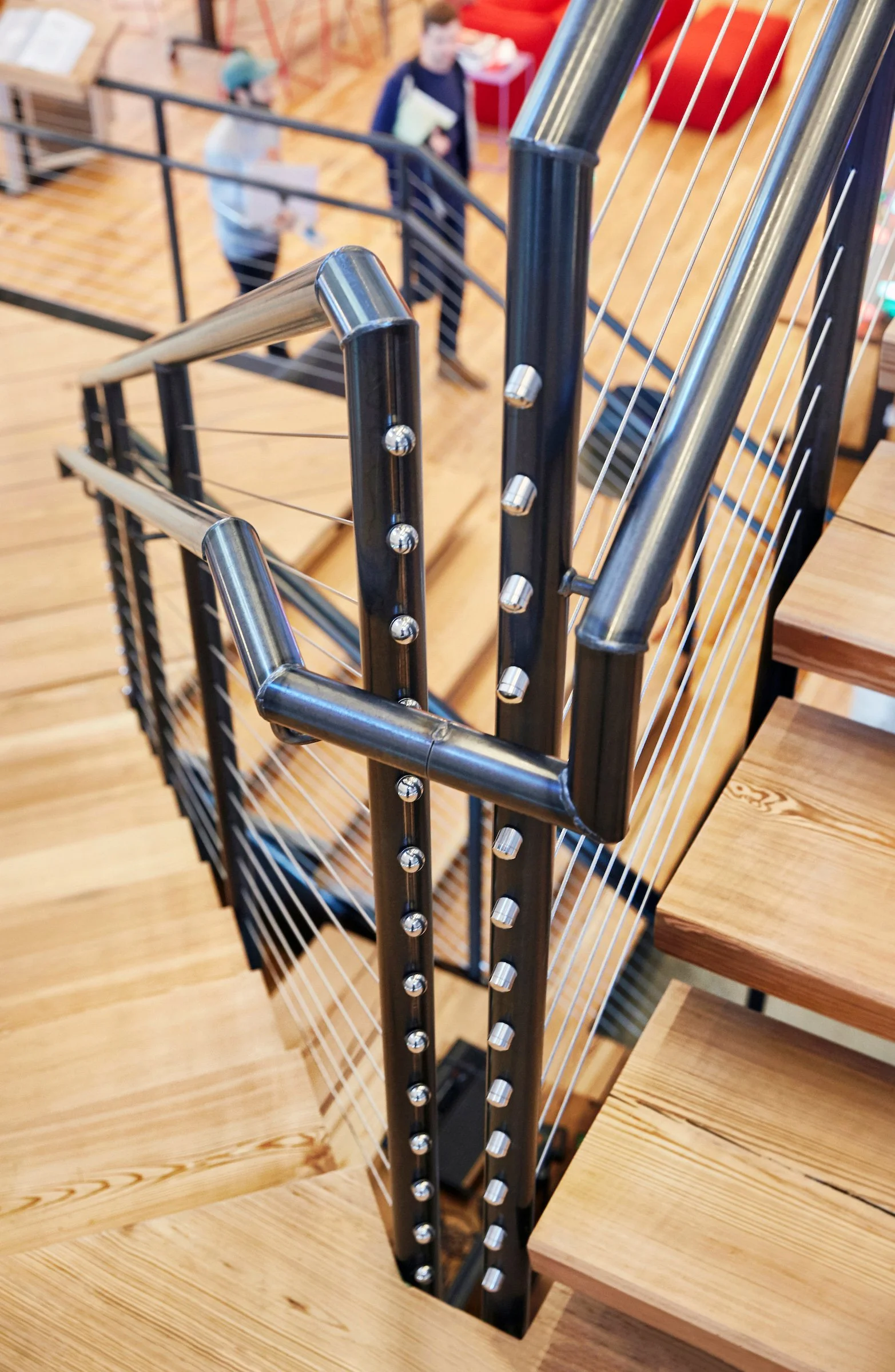 Close-up of a staircase railing with metal handrails and thin cables, showing wooden stairs and a blurred view of people in the background.