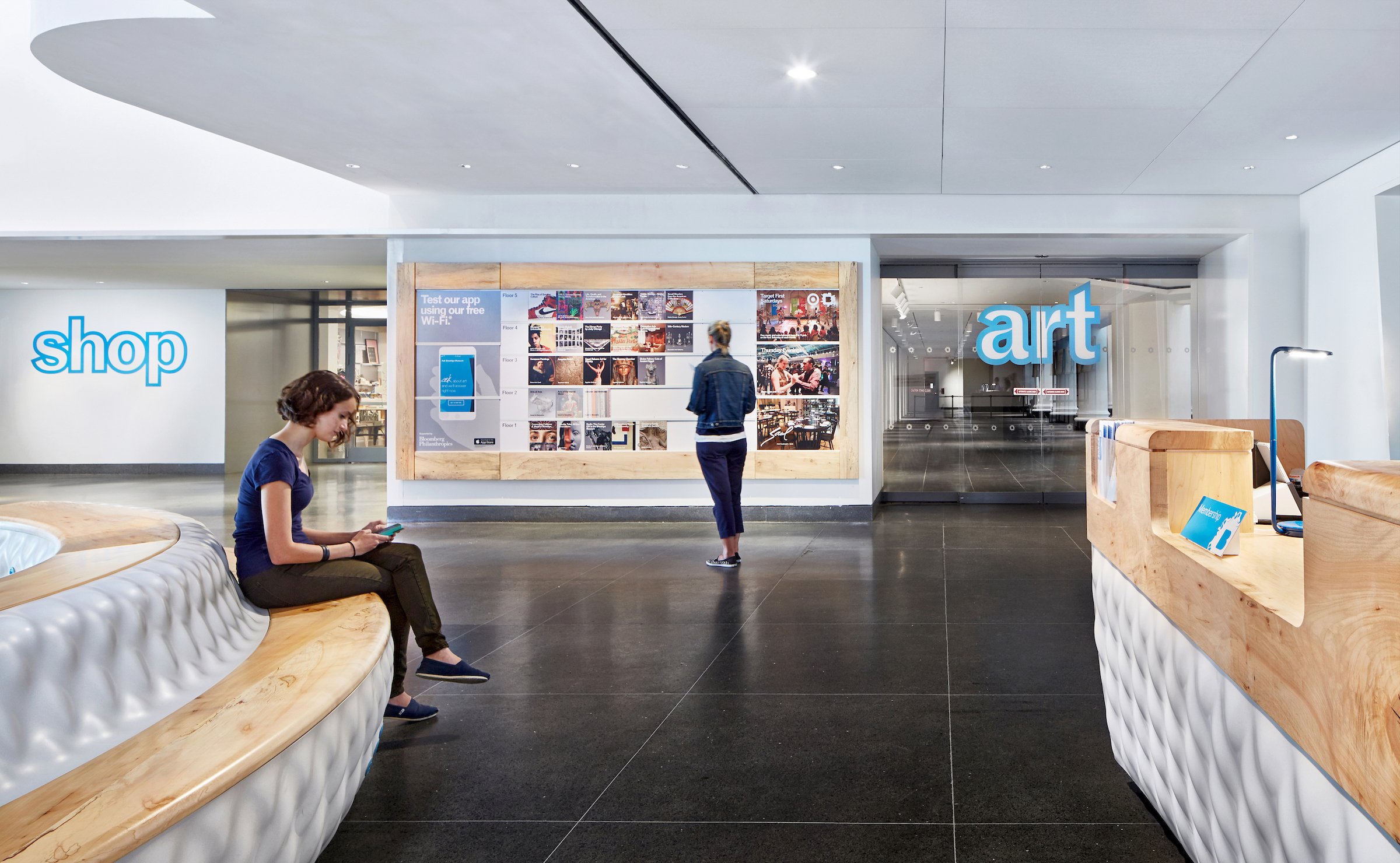 Interior of an art gallery or museum with the words 'shop' and 'art' on the walls, a woman sitting on a curved bench using her phone, and another woman looking at artwork on the wall.