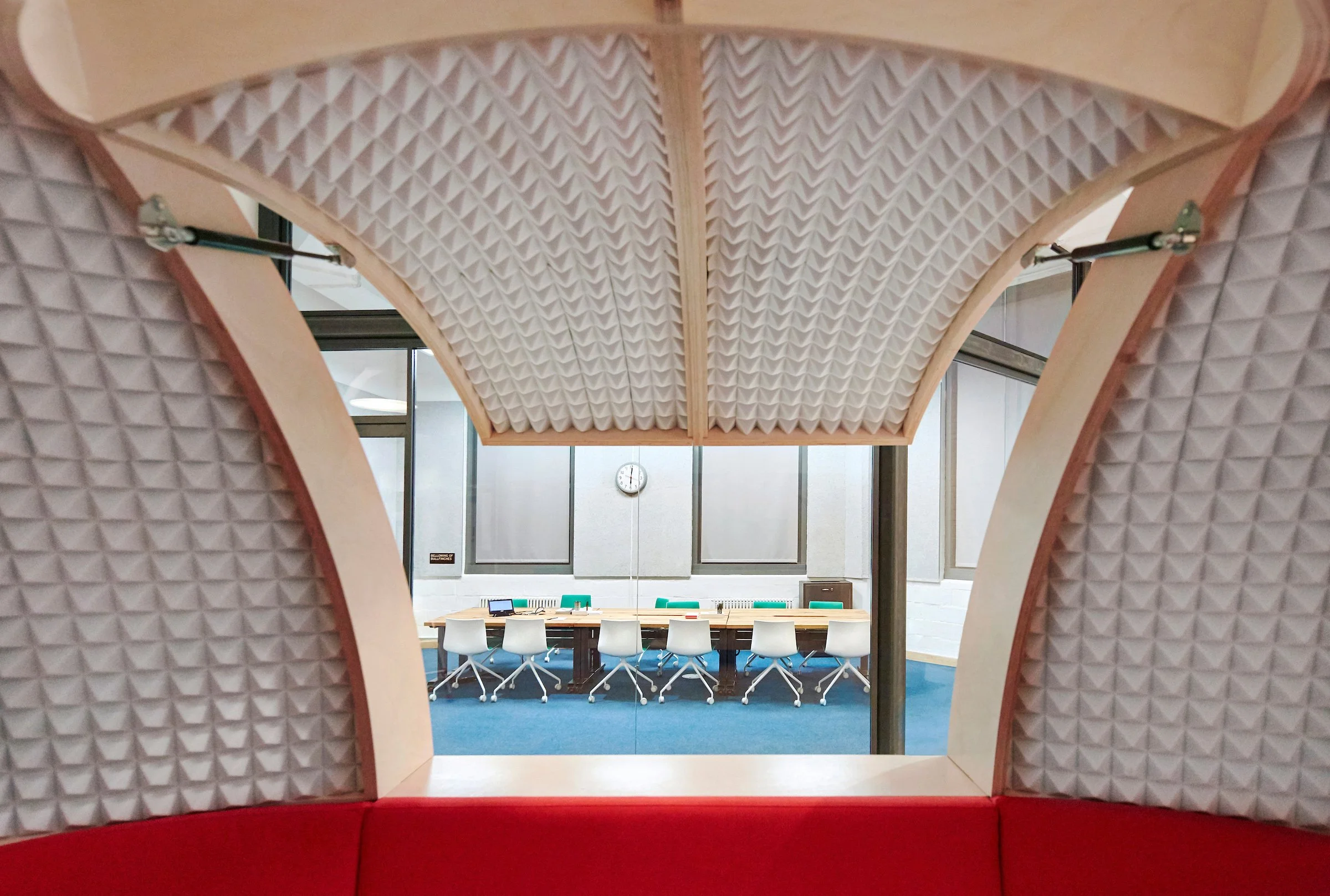 A conference room with a long table and white chairs, seen through a soundproofed panel with padded, textured walls, and a window showing the room and a clock on the wall.