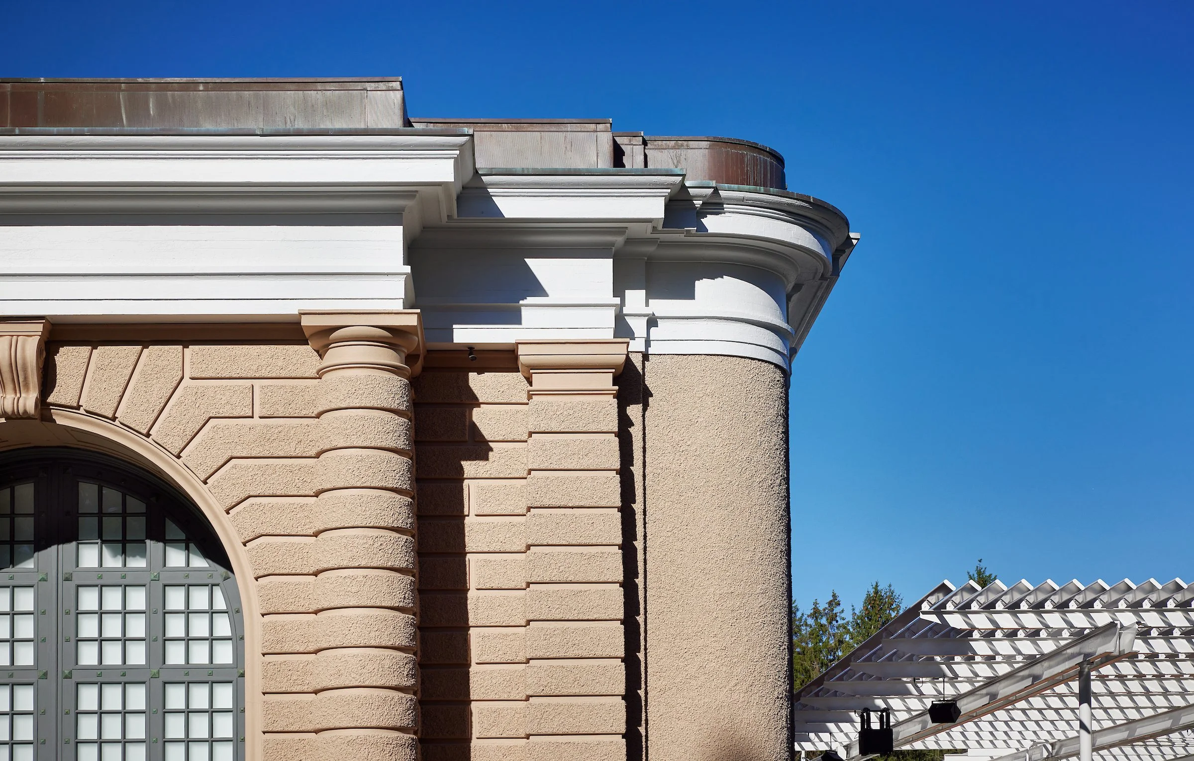 Close-up of a building corner showing beige stone and stucco walls, a large arched window, and a white decorative cornice under a clear blue sky.