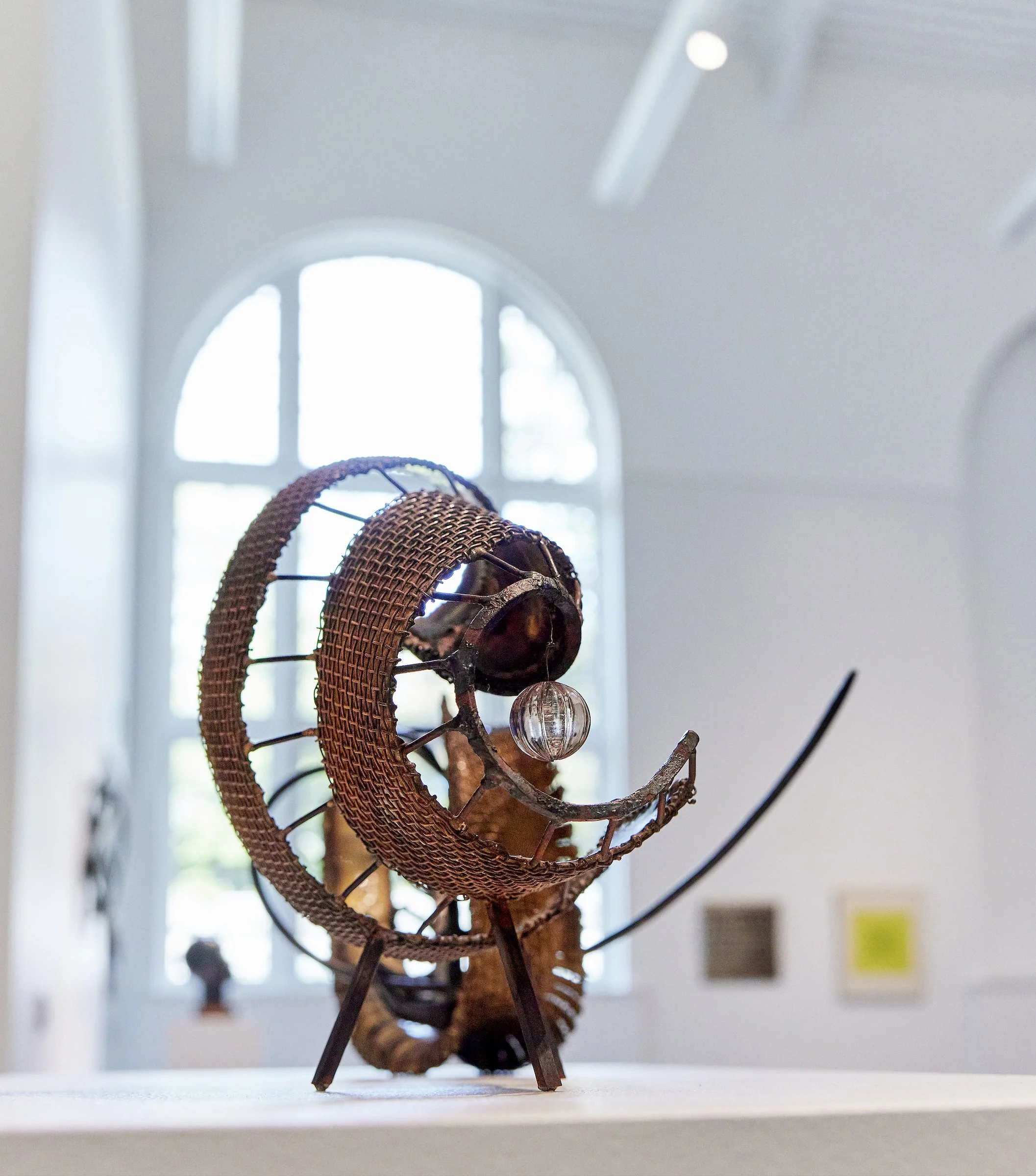 A modern art sculpture resembling a chameleon, made of metallic and woven materials, with a glass or crystal orb in its mouth, displayed on a white pedestal in a gallery with large arched windows.