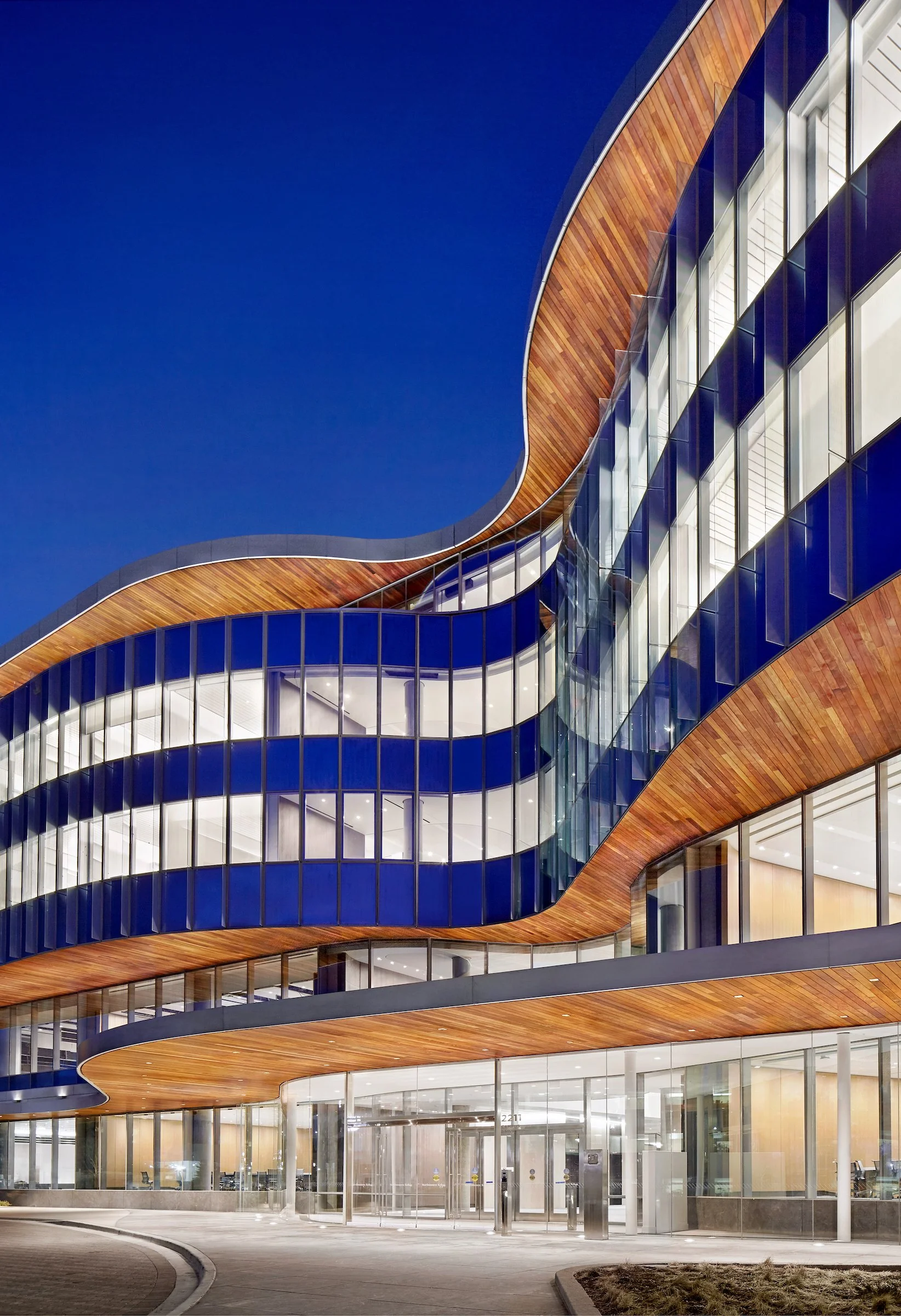 Modern building with curved glass and wood accents illuminated at night, featuring multiple floors and large glass windows.