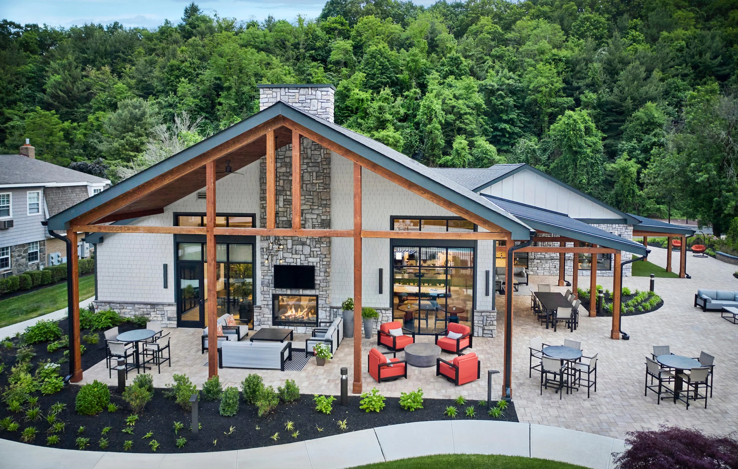 A modern house with white and stone exterior, large windows, and a covered patio with outdoor furniture, a fireplace, and a view of green hills and trees in the background.