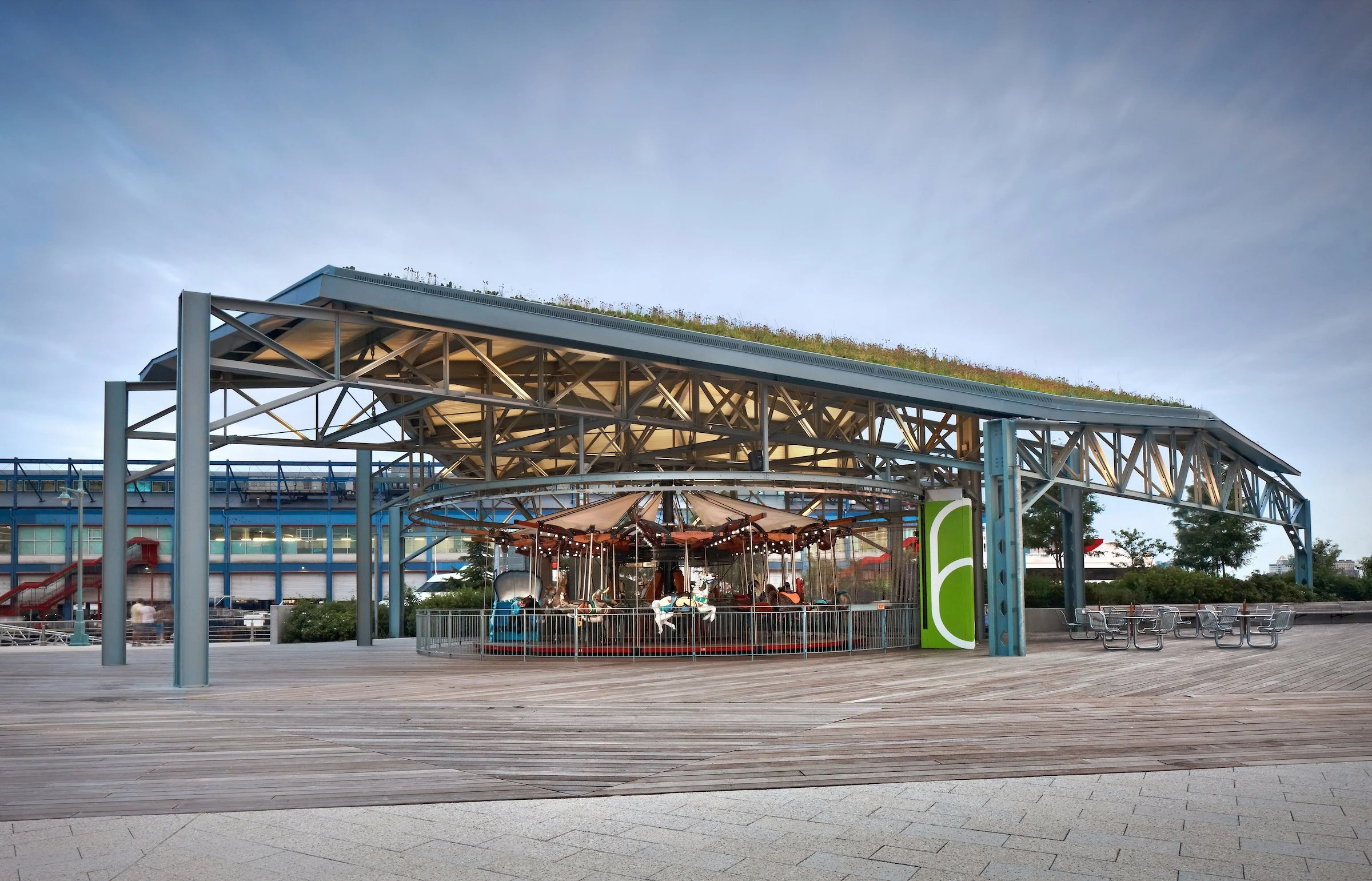 An outdoor carousel ride located under a modern steel canopy with a green rooftop, surrounded by empty benches and a wooden promenade.