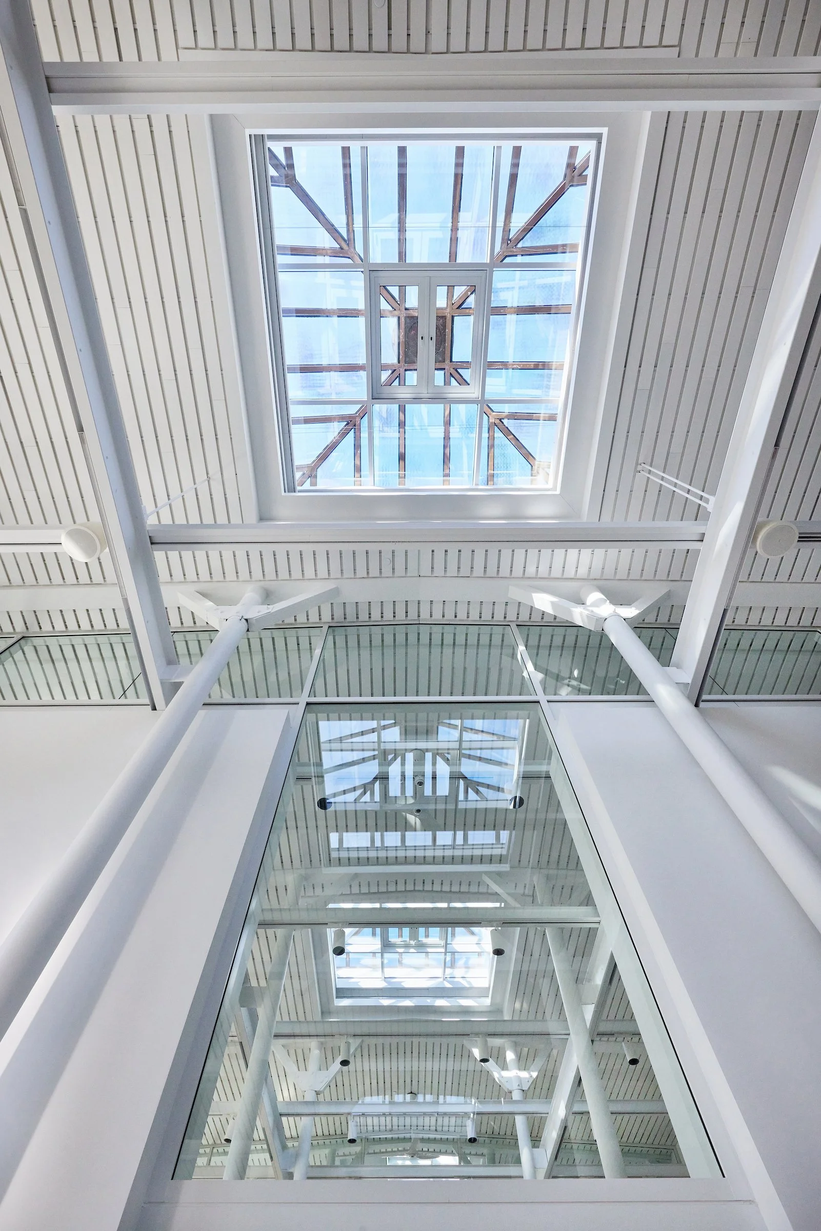 Looking up at a glass ceiling with a skylight framed by white beams, with reflections visible in glass windows and white structural supports below.