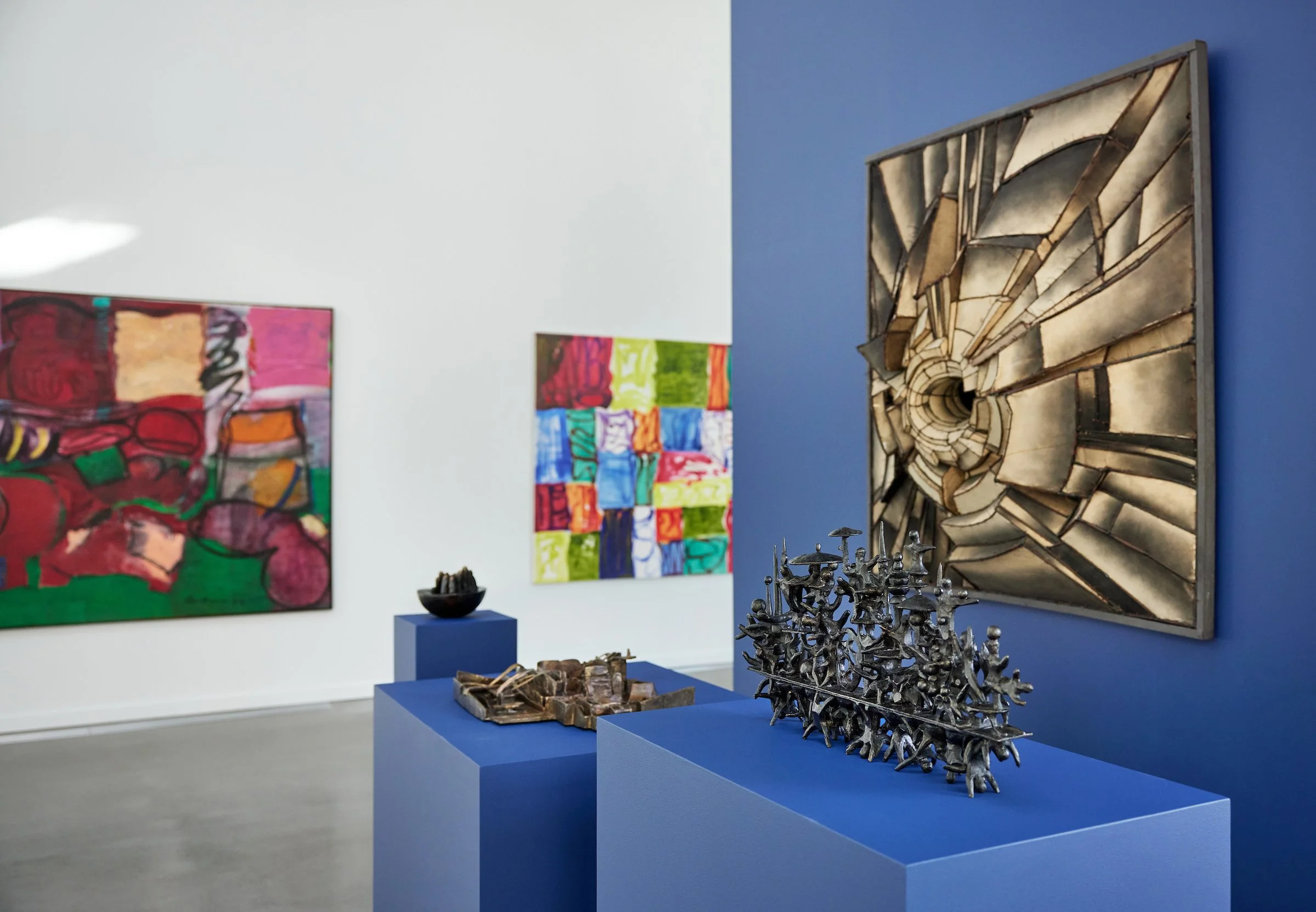 Art gallery featuring colorful abstract paintings on white walls, with metal sculptures on blue pedestals in the foreground, and a geometric metal wall sculpture on a blue wall.