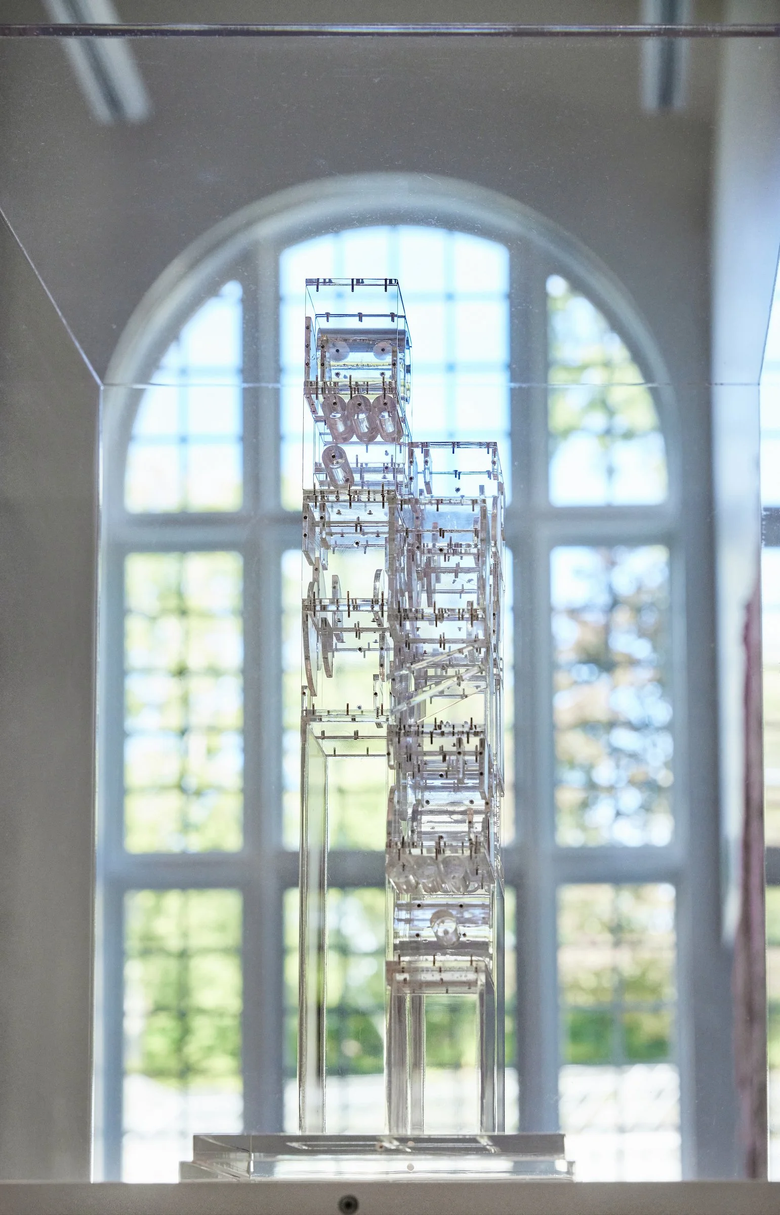 A transparent sculpture made of interconnected clear acrylic or glass elements, displayed in front of a large arched window with grid panes, showing trees and blue sky outside.