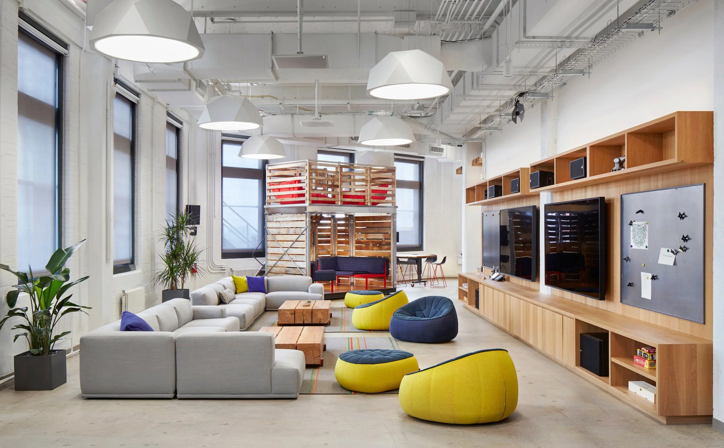 A modern open office lounge with large windows, white walls, and ceiling lights. There are assorted sofas and chairs, a coffee table, indoor plants, and a wooden entertainment unit with two large screens. In the background, there is a lofted area wit