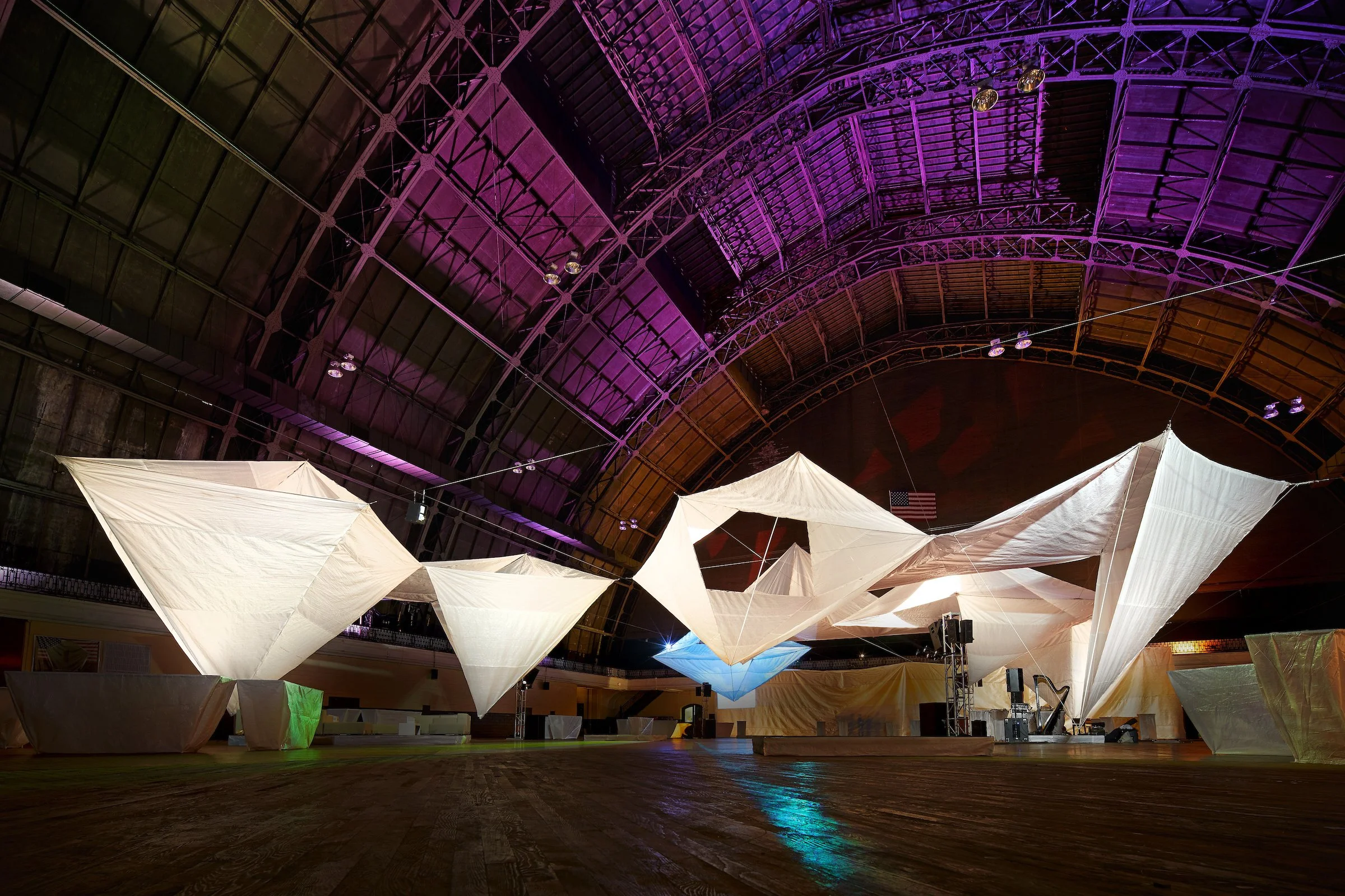 An indoor event space with large, abstract white fabric installations suspended from the ceiling, illuminated with colorful lighting in purple, blue, and green hues.
