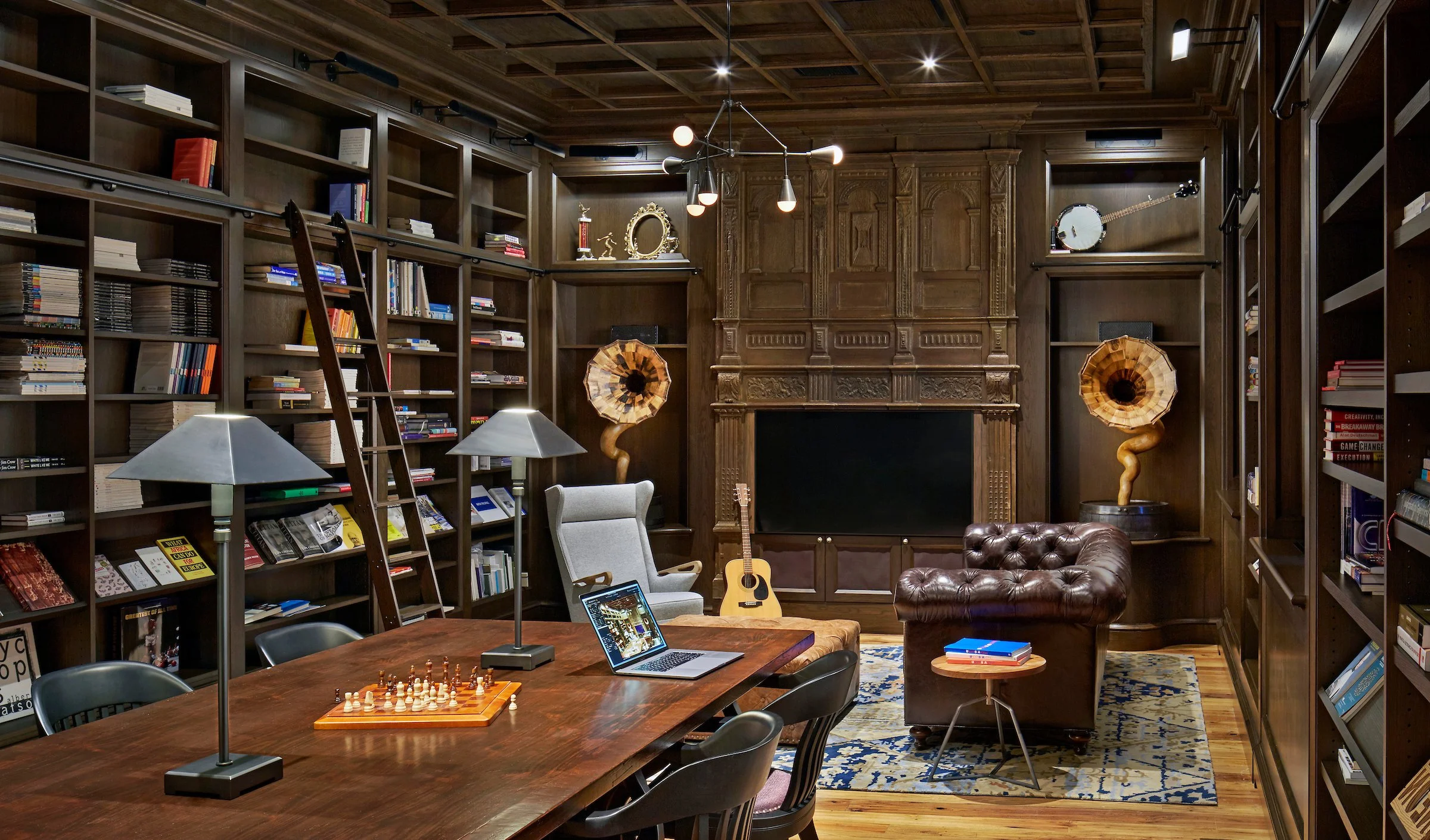 A spacious room with dark wooden bookshelves filling the walls, filled with books and decorative items. In the center, there is a wooden table with two lamps, a chessboard, and chairs around it. Against the back wall, there is a gray armchair, an aco