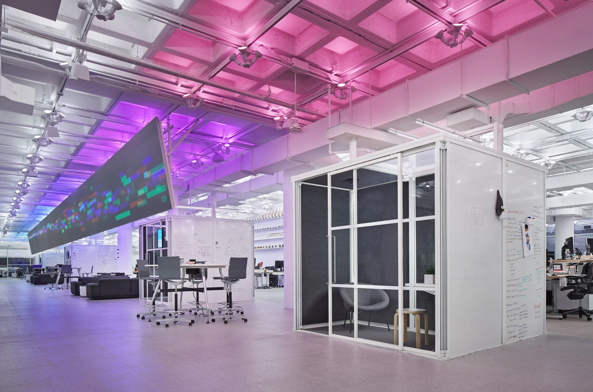 Modern open-plan office space with a white cubicle, whiteboard walls, office chairs, and a large digital display hanging from a colorful illuminated ceiling.