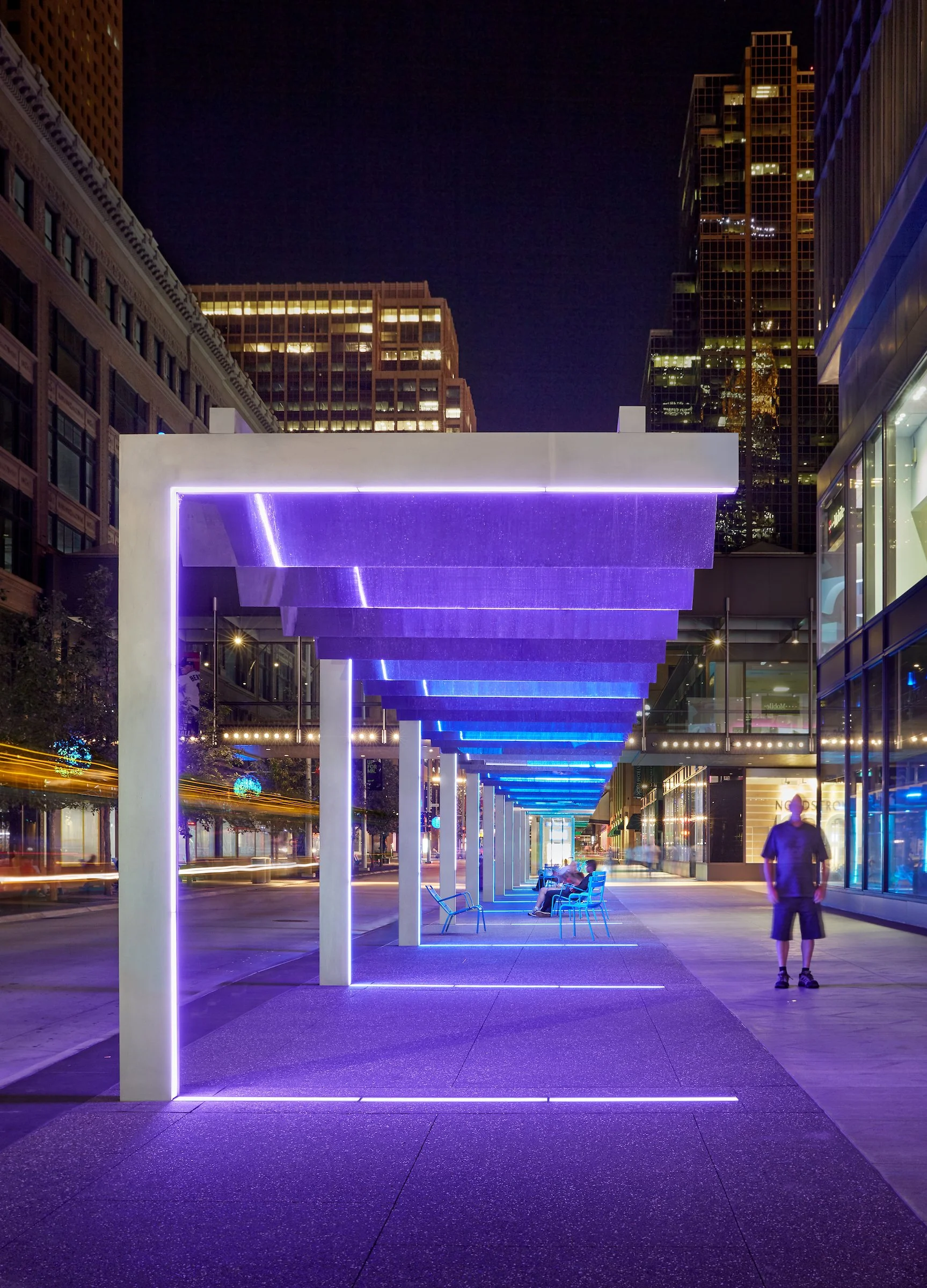 Night view of a modern city sidewalk with illuminated purple-blue canopy and benches, with tall buildings in the background and a person walking on the sidewalk.