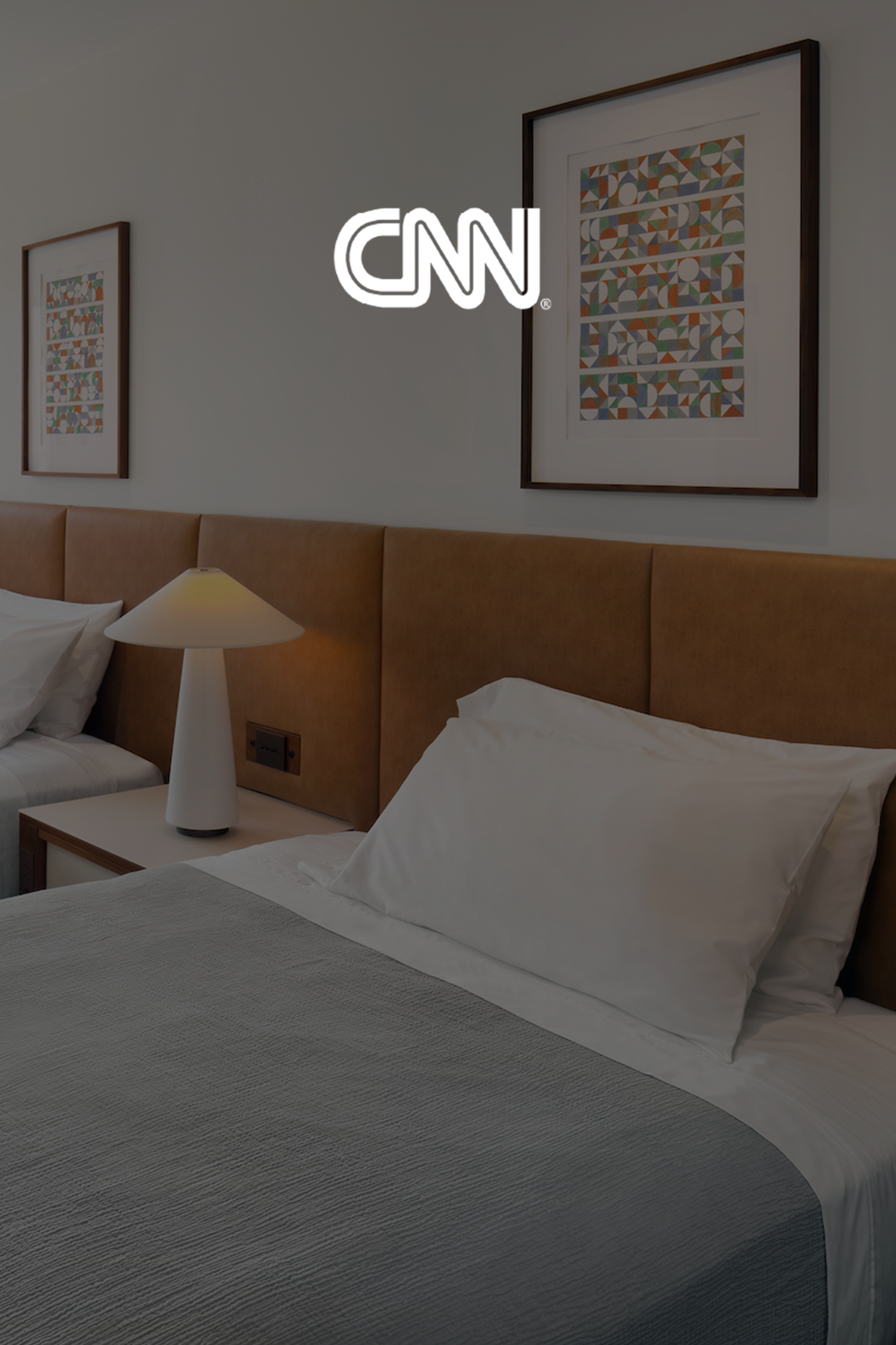 Cover photo of CNN website featuring a hotel room with a bed, white pillows, a bedside table with a lamp, and framed artwork on the wall.