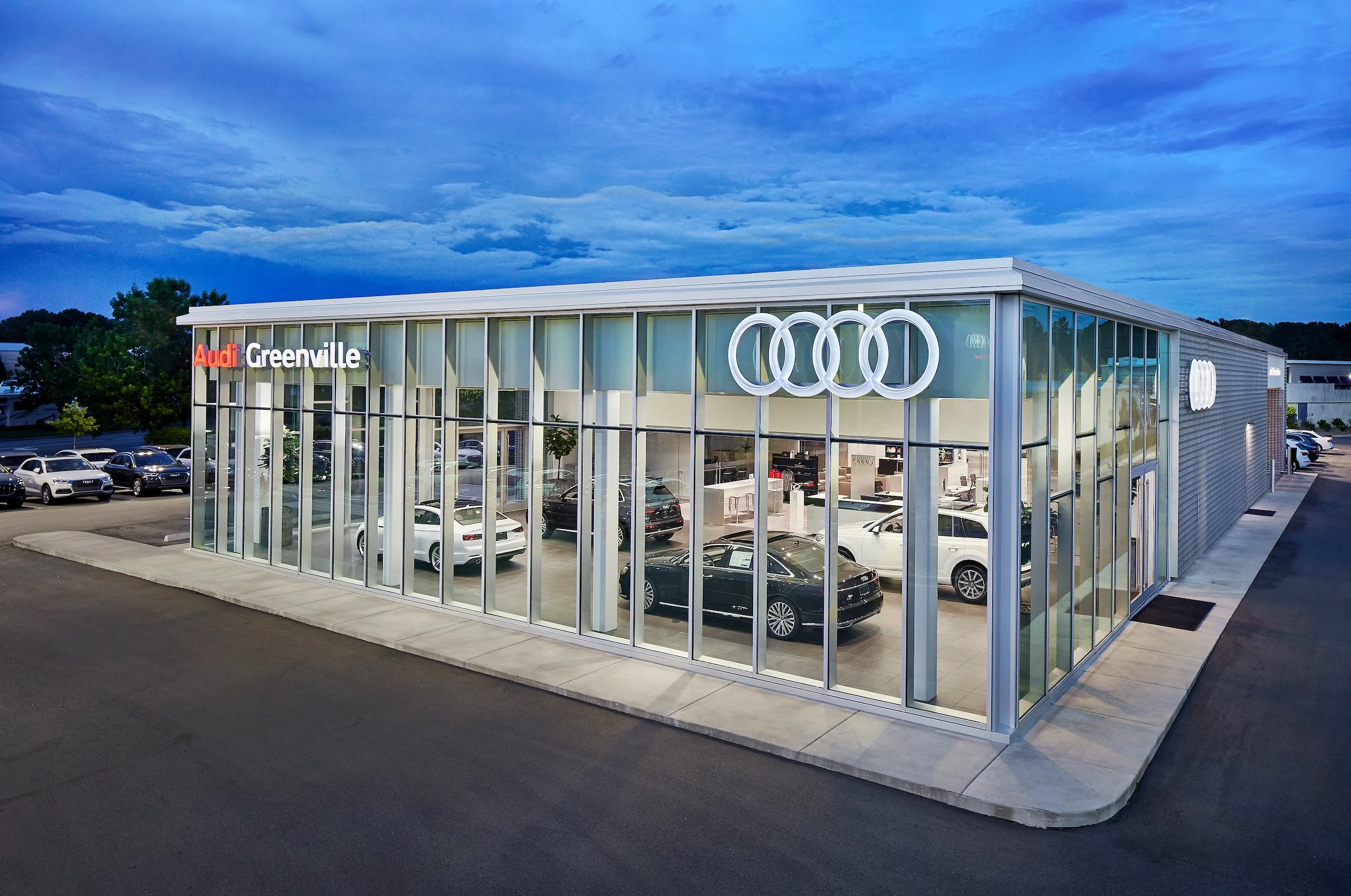 Exterior view of an Audi dealership with large glass windows displaying cars inside, signage with Audi logo, and the location 'Greenville' on the building.