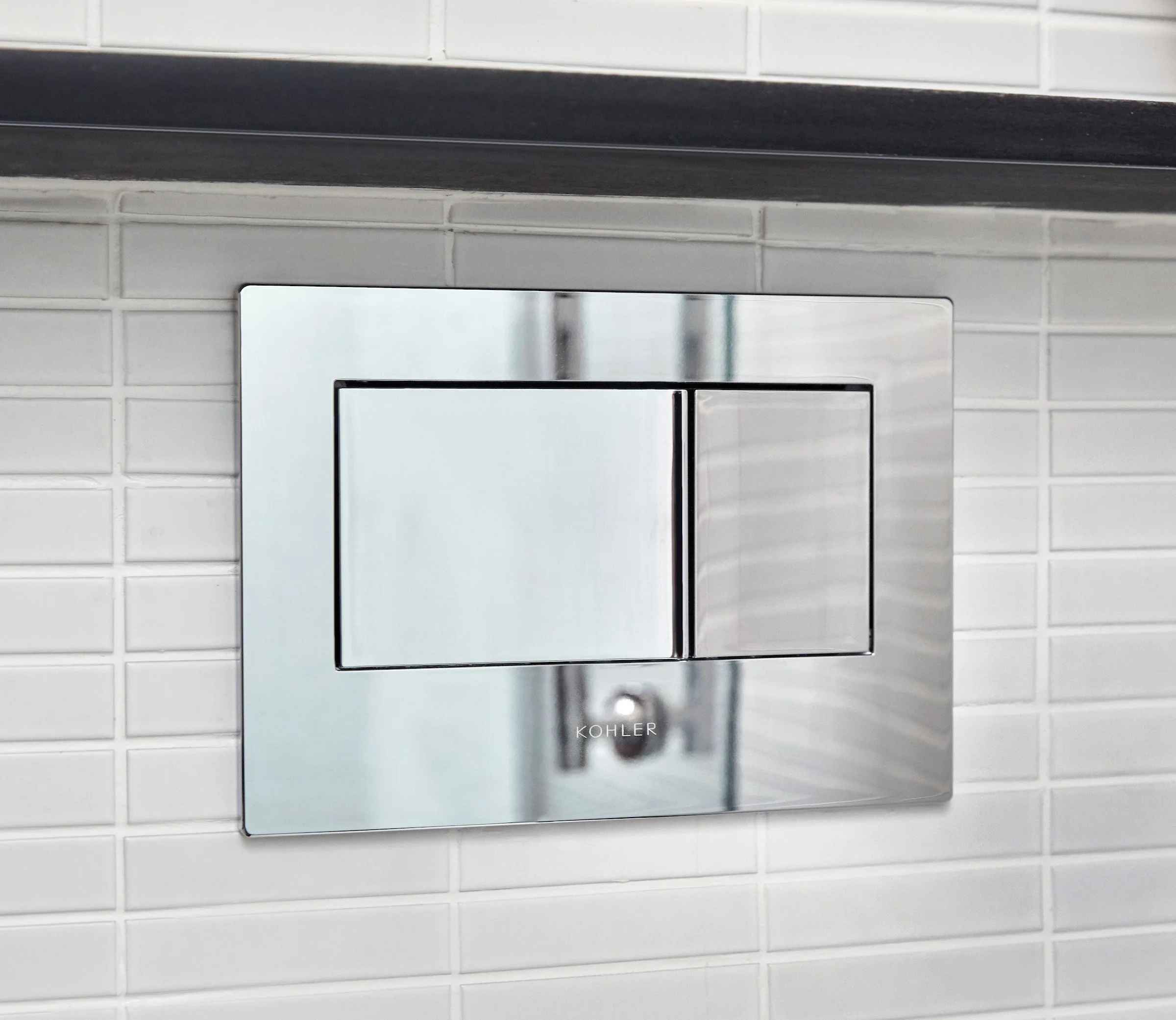 Close-up of a modern flush panel for a toilet, mounted on a wall with white subway tiles, with the brand name 'Kohler' visible on the panel.