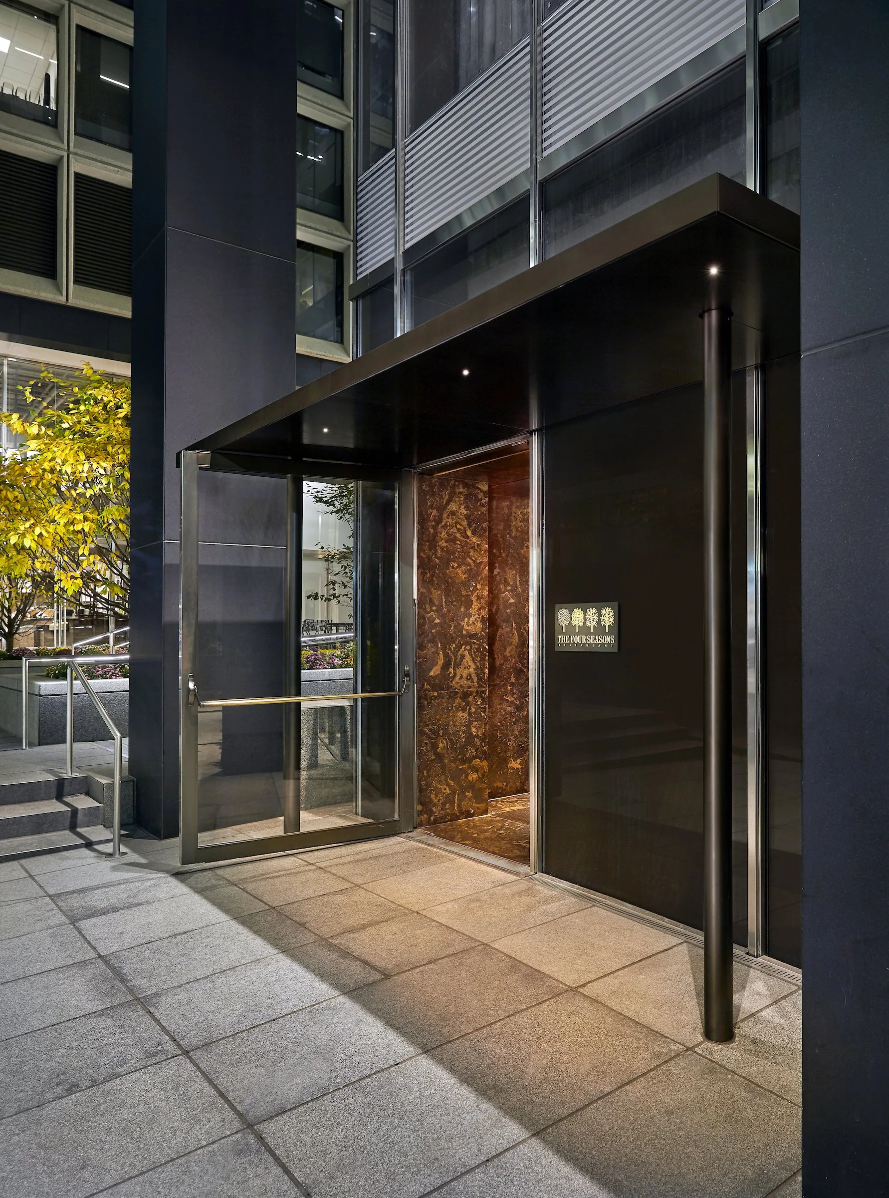 Modern building entrance with a glass door, black and brown marble walls, metal railing, and an illuminated sign that reads 'The Four Seasons' with a tree logo.
