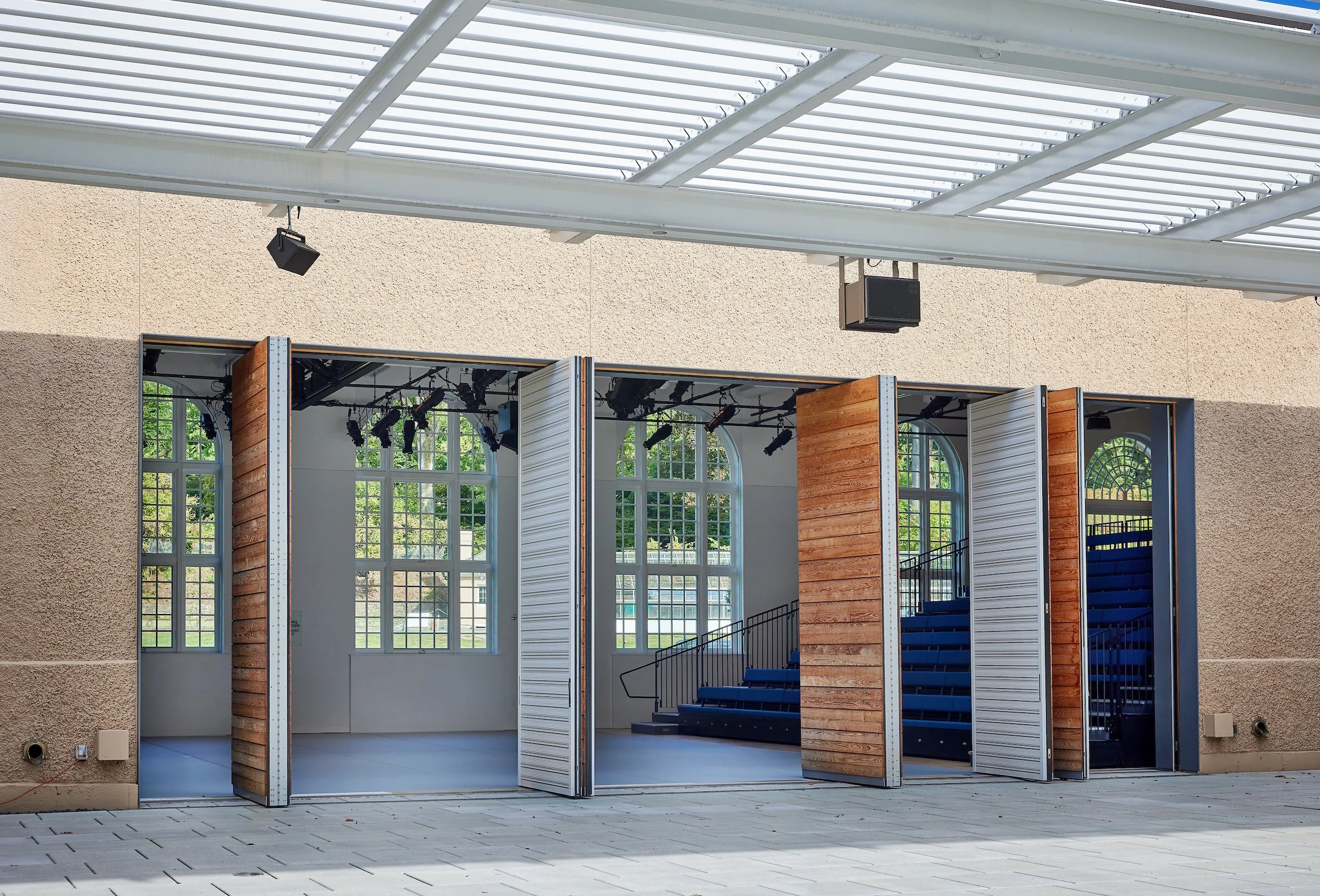 Indoor stage with open folding partitions, large arched windows, blue seating, and stage lighting equipment.