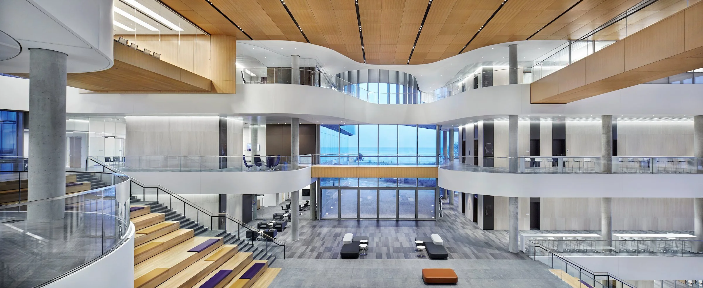 Modern, spacious lobby with multiple levels, glass railings, wooden accents, and large windows providing natural light and a view outside.