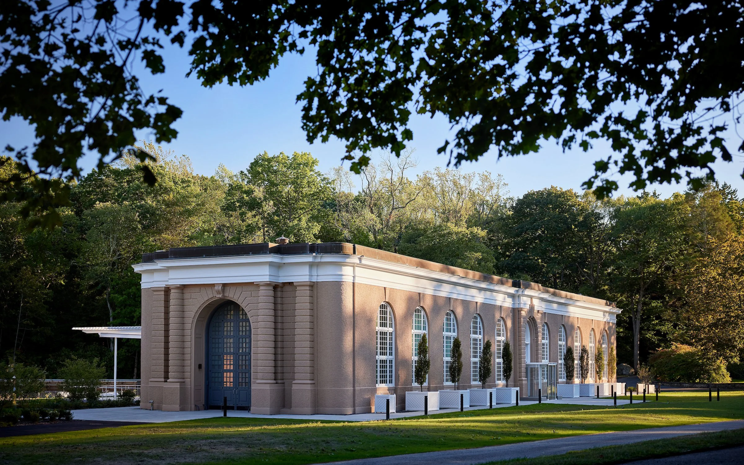 A historic brick building with large arched windows, white trim, and a blue door, surrounded by green trees and grass.