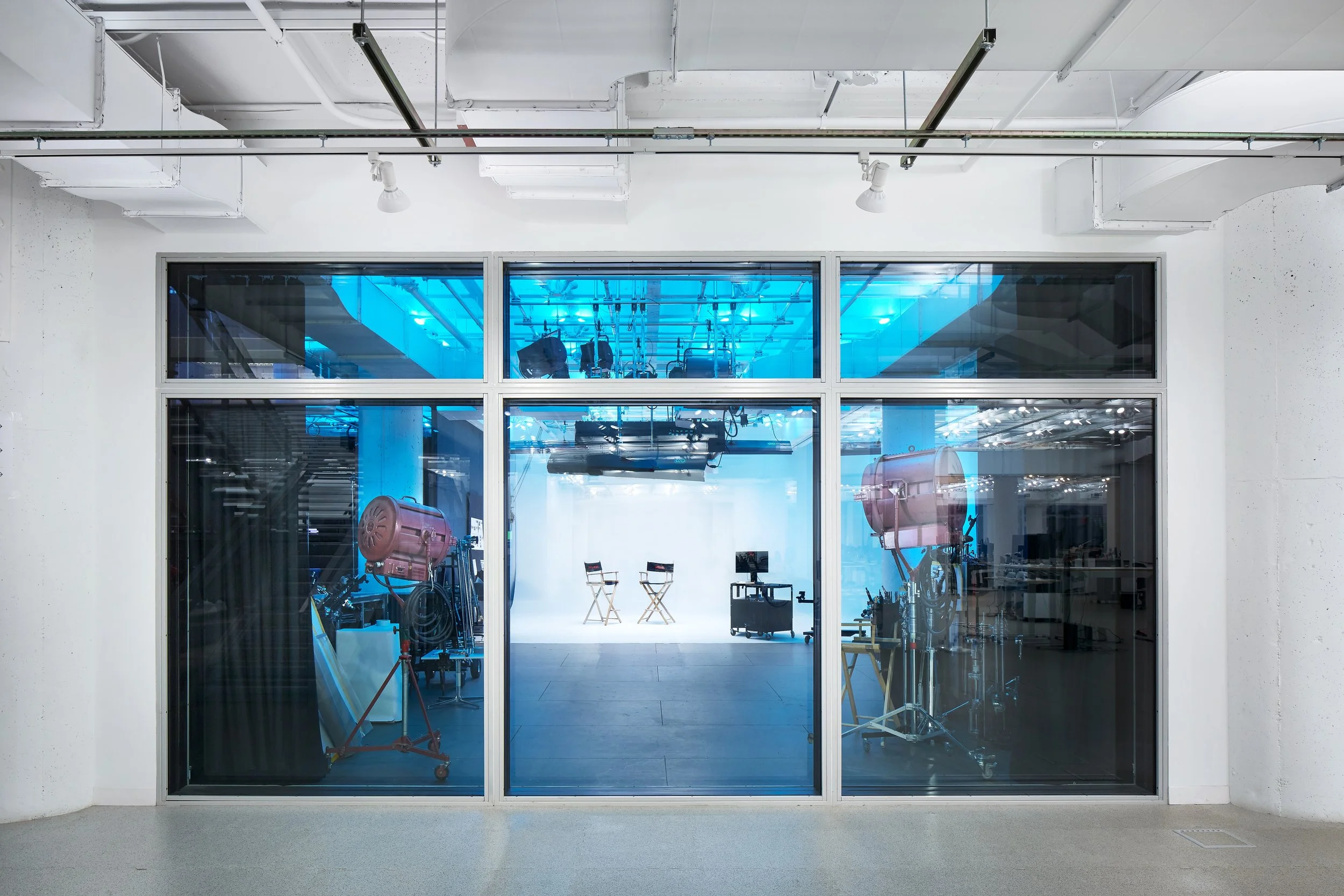 View through glass wall showing a radio or TV studio with two director chairs, studio lights, and equipment inside.
