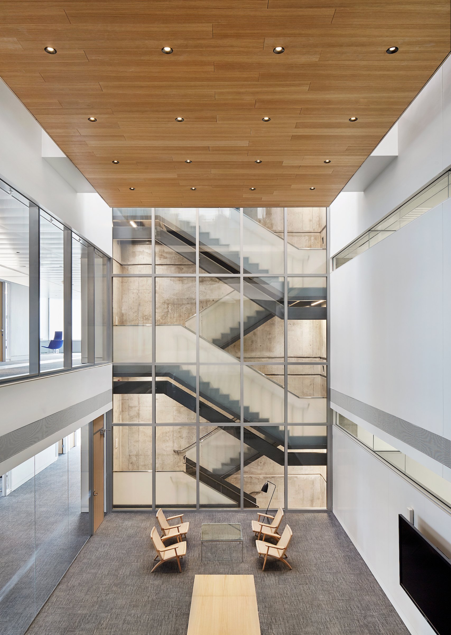 Modern interior lobby with a high ceiling, wooden panels, and small recessed lights. A large glass staircase with metal framing is visible behind a glass wall, and there is a sitting area with four beige chairs around a small table on a gray carpeted
