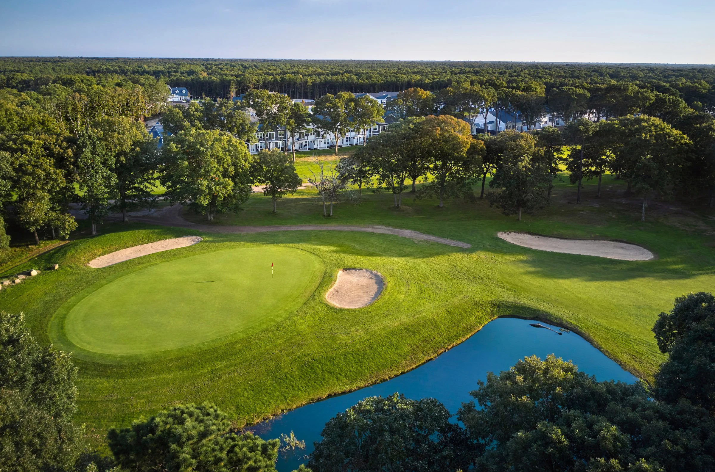A golf course with green grass, sand bunkers, a small pond, trees, and a residential neighborhood in the background under a clear sky.