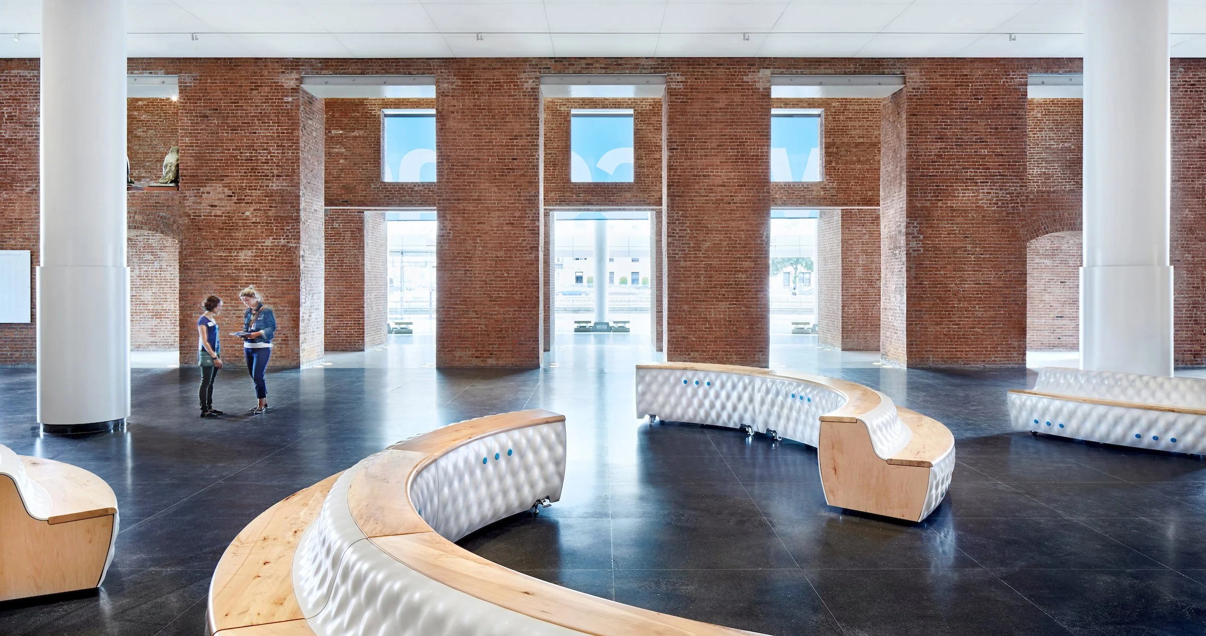 Interior of a modern building with brick walls, large windows, circular white and wood benches, and two women standing and talking in the spacious area.